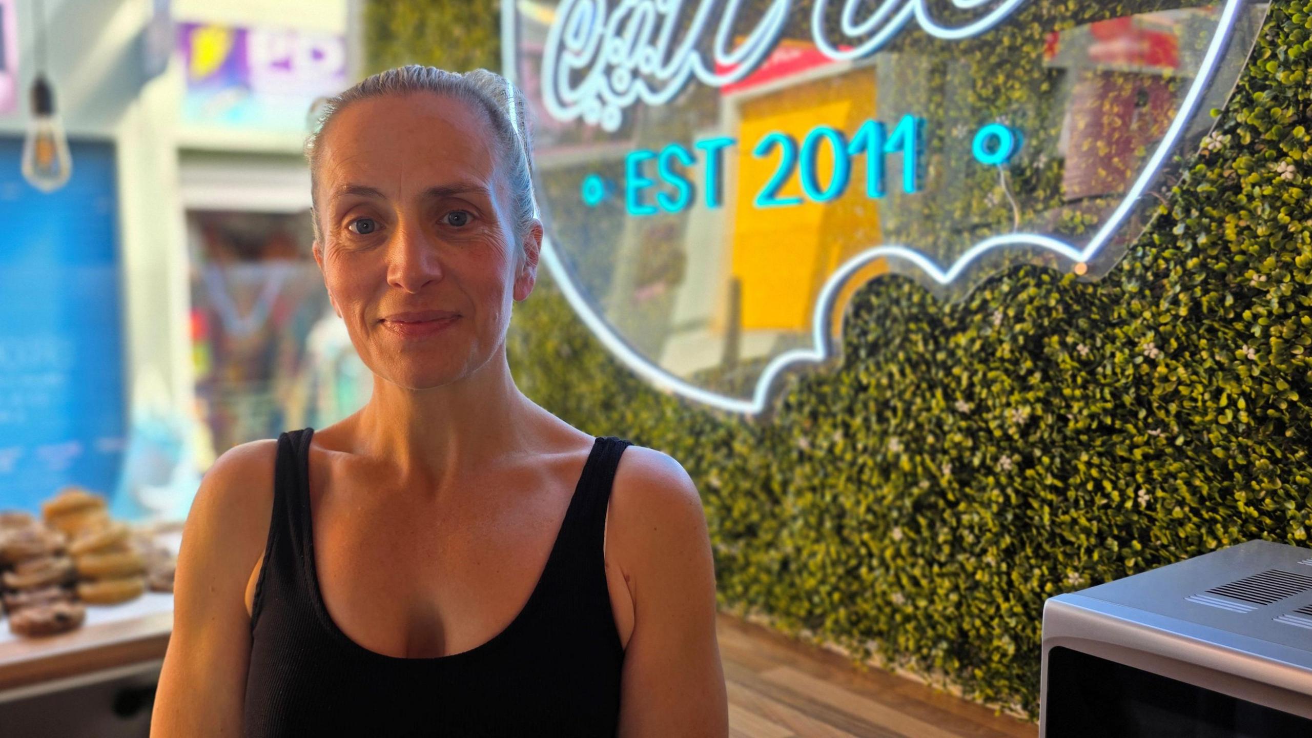 Tori Fama is wearing a black vest-type top. Behind her is a neon sign where we can see "Est 2011" - and fake grass on the wall.
