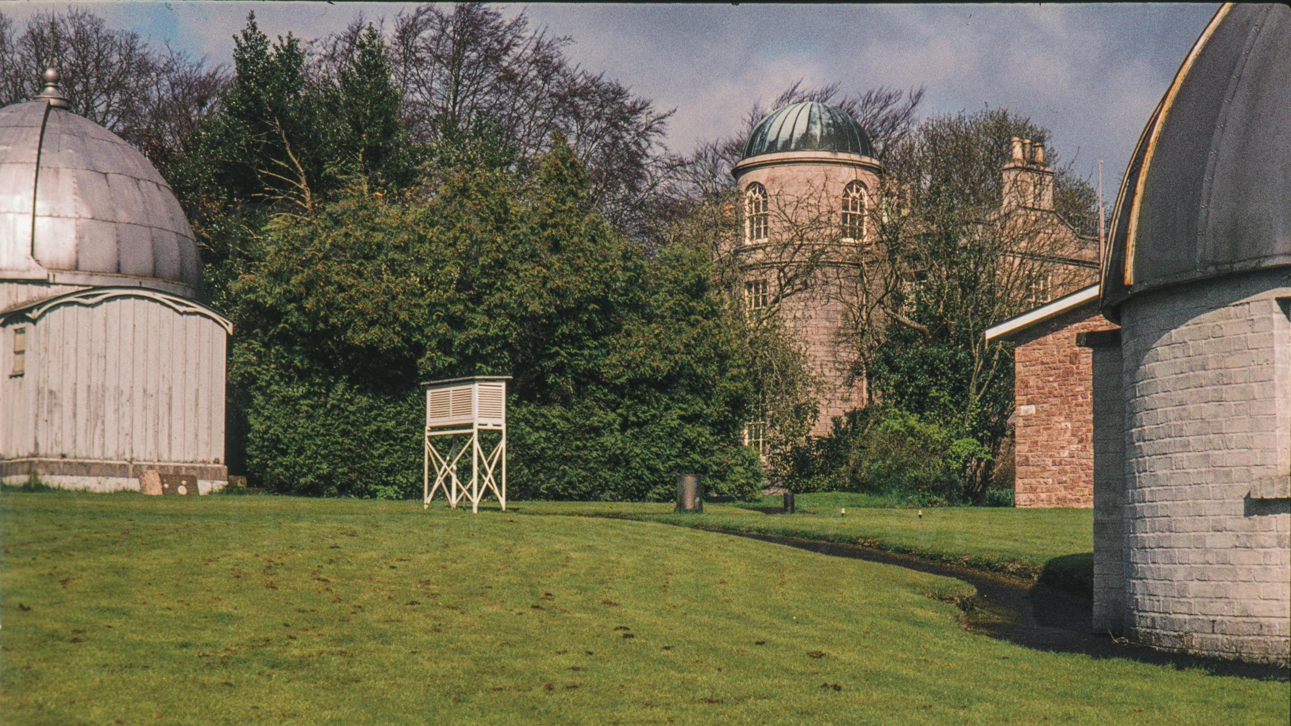 A digitalised photo from a film of sphere buildings made of metal. South view of the observatory looking over the lawn with the dome of the Grubb 10 inch refractor on the left the weather station Stevenson Screen centre and the main observatory and library building on the right.