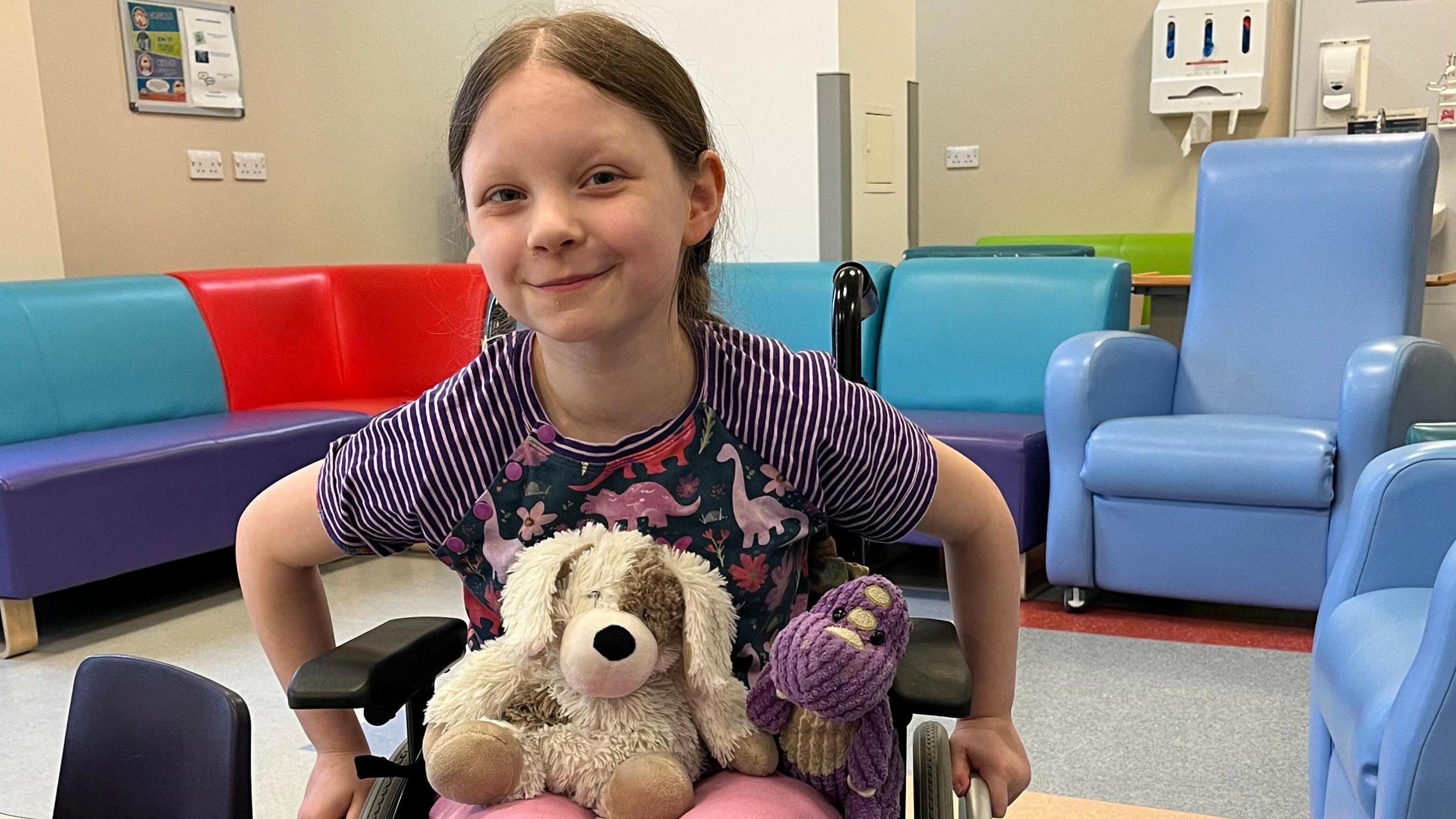 Ivy is sitting in a wheelchair in what looks like a hospital waiting area and smiling at the camera. Two teddy bears sit on her lap. Behind her is a multi-coloured L-shaped vinyl sofa and a couple of blue very high-back armchairs.