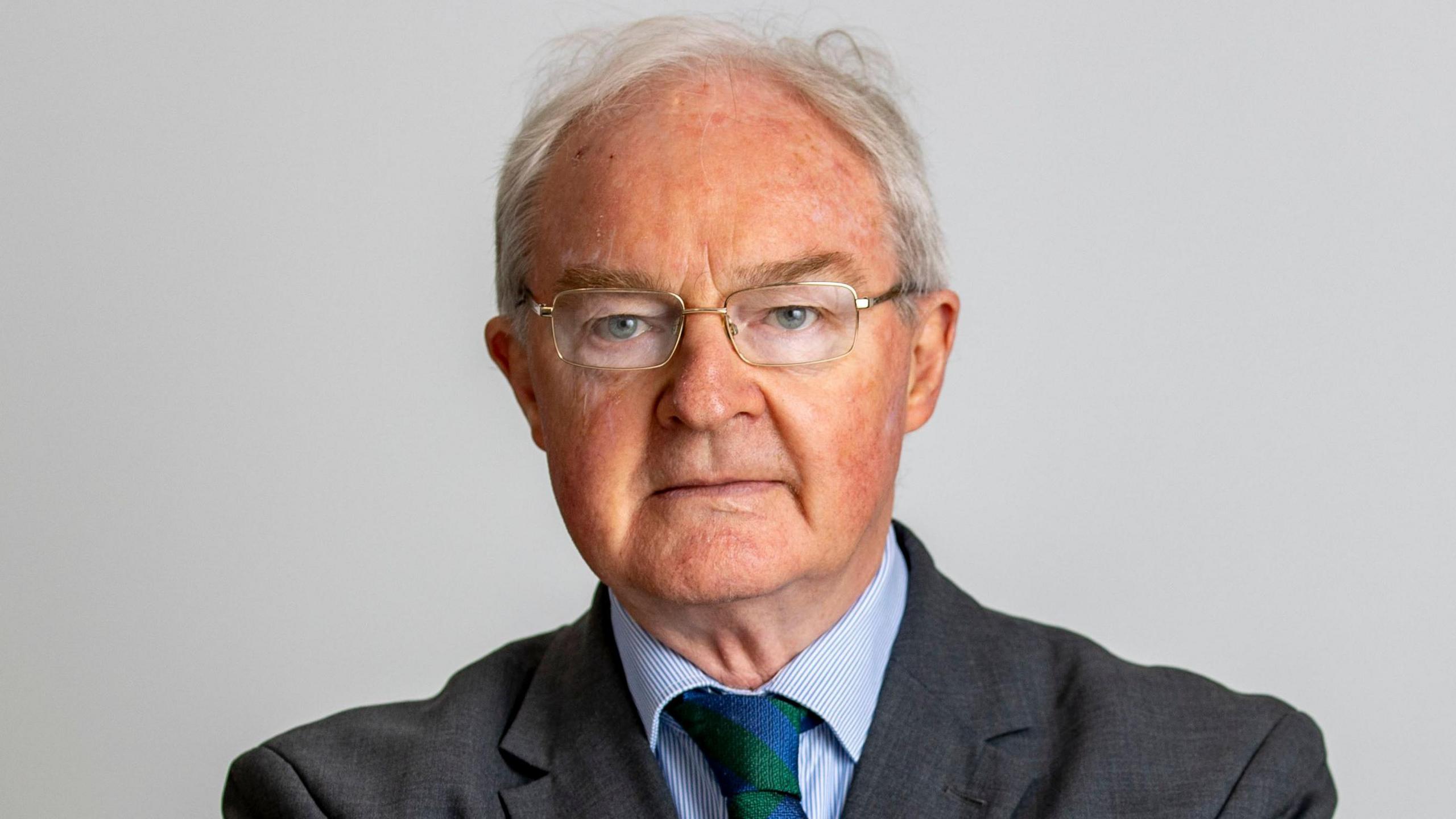 Sir Declan Morgan looking at the camera, wearing glasses, a dark suit, a blue shirt and a striped tie