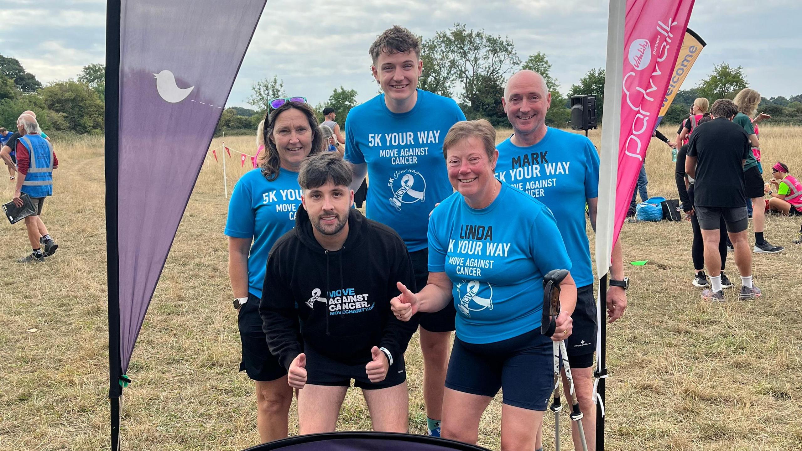 Three men and two women posing for a photo in between flag posts at a park run. Four of them are in blue Move Against Cancer t-shirts and Adam is at the front with both thumbs up, wearing a black Move Against Cancer hoody. There are people in the background talking and sitting.
