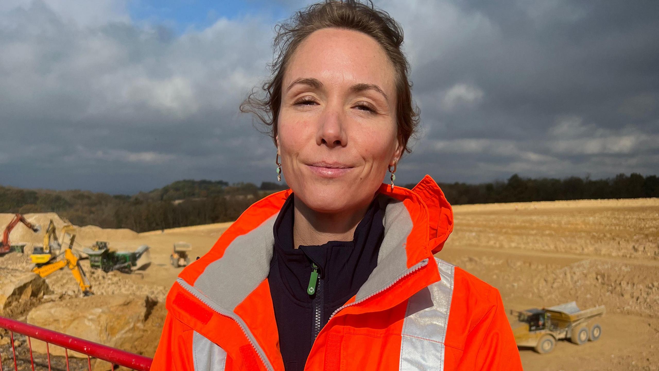 Celine Acard, with hair tied up, wearing an orange hi vis jacket standing in front of the construction site