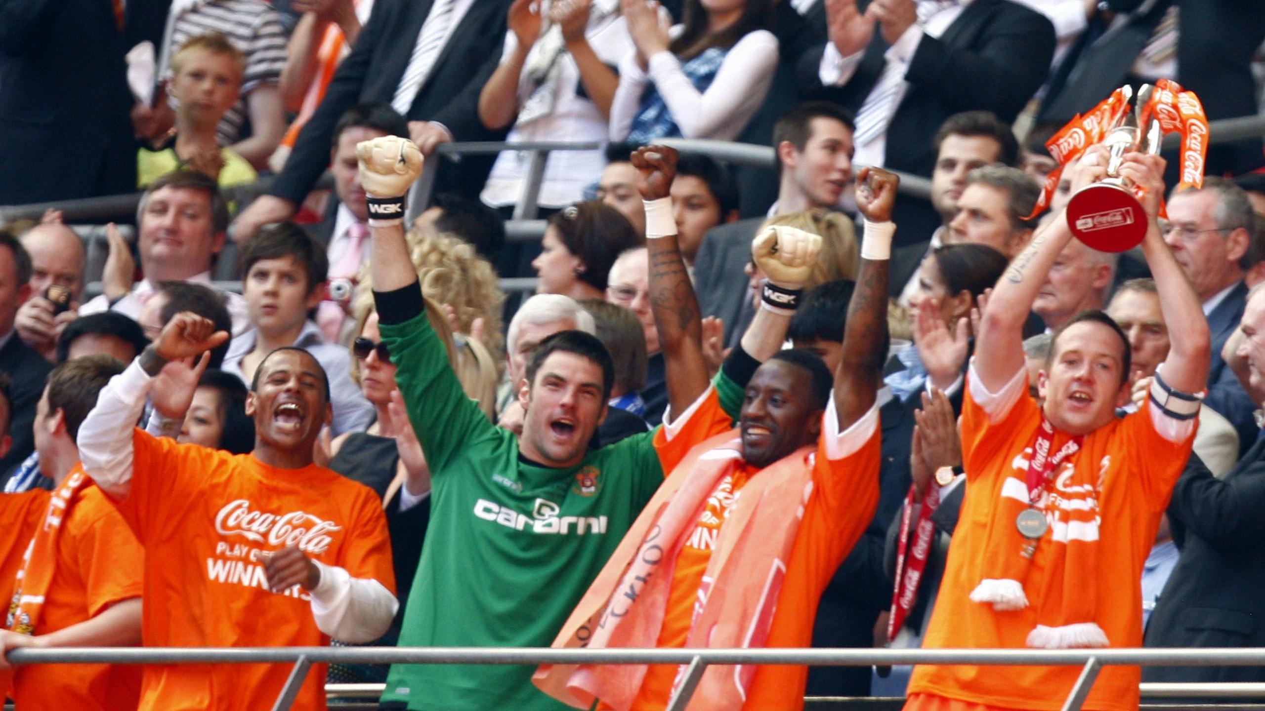 Charlie Adam of Blackpool in an orange shirt lifts the play-off trophy above his head while his team-mates raise their arms in celebration