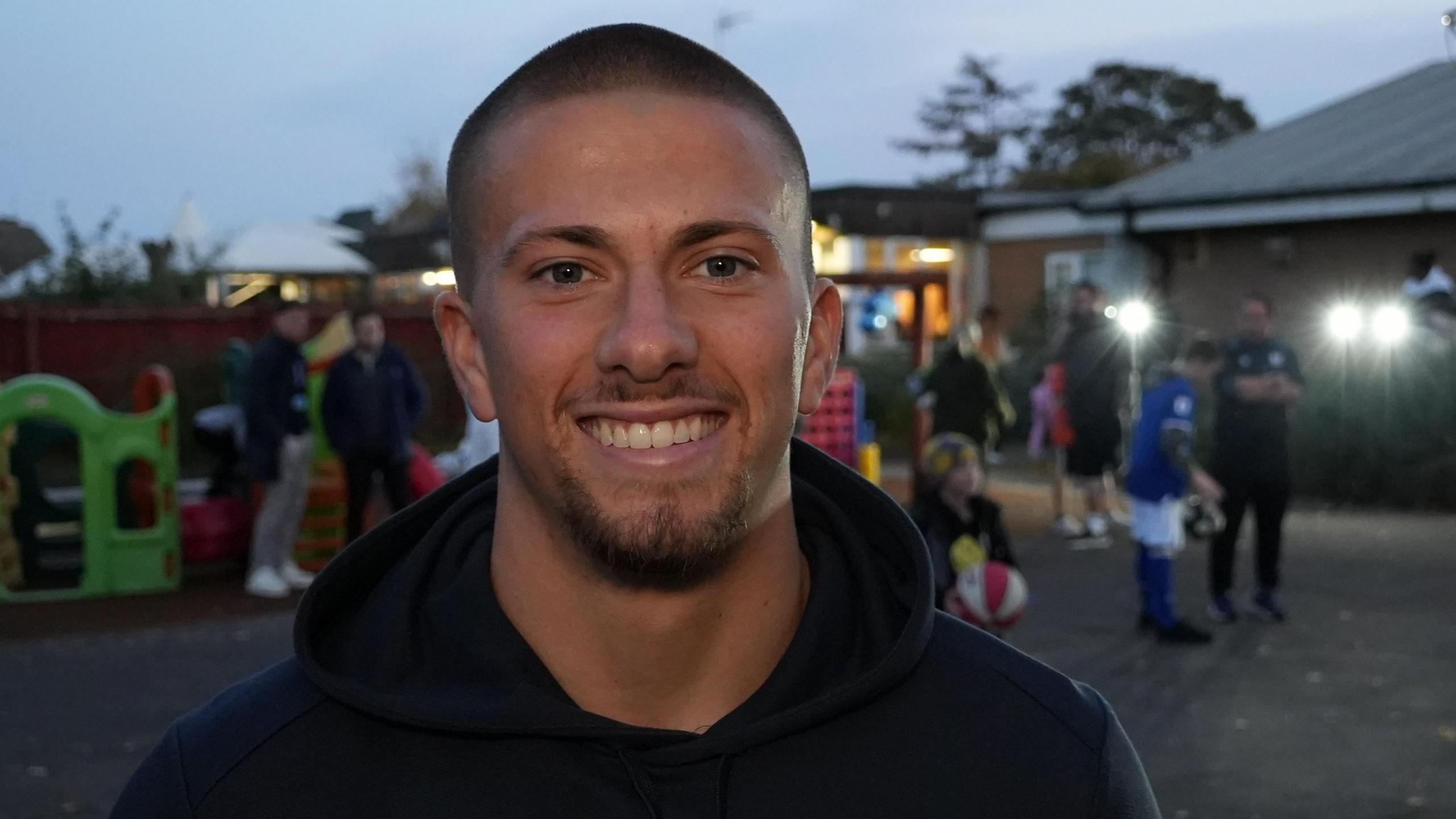 Harry Clarke smiles at the camera outside a family hub as it begins to get dark outside. Children and adults can be seen enjoying a playground behind him. He wears a black hoodie.