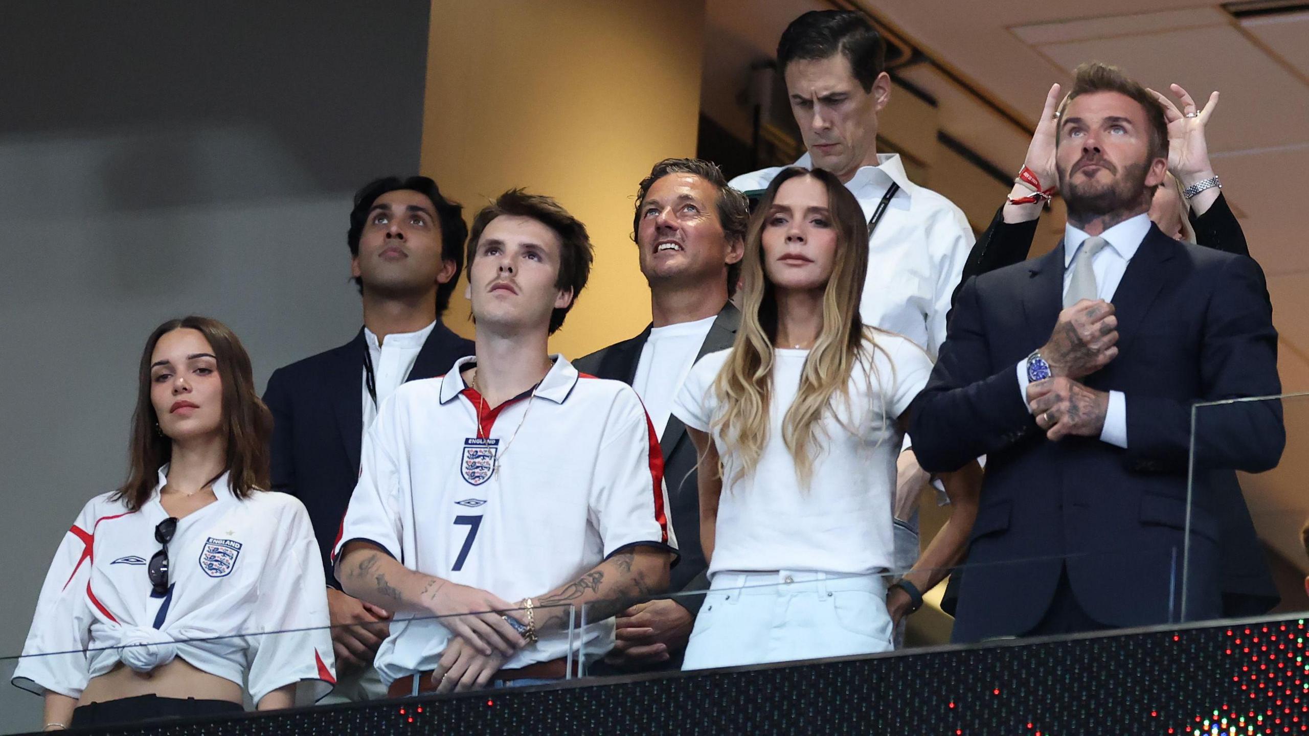 A picture of Jackie Apostel, Cruz Beckham, Victoria Beckham, David Beckham and Ken Paves in a box at a football match during the World Cup between England and Argentina. The group, dressed in a mixture of England shits and formal clothing, appear to be looking up at a screen, which is out of shot.