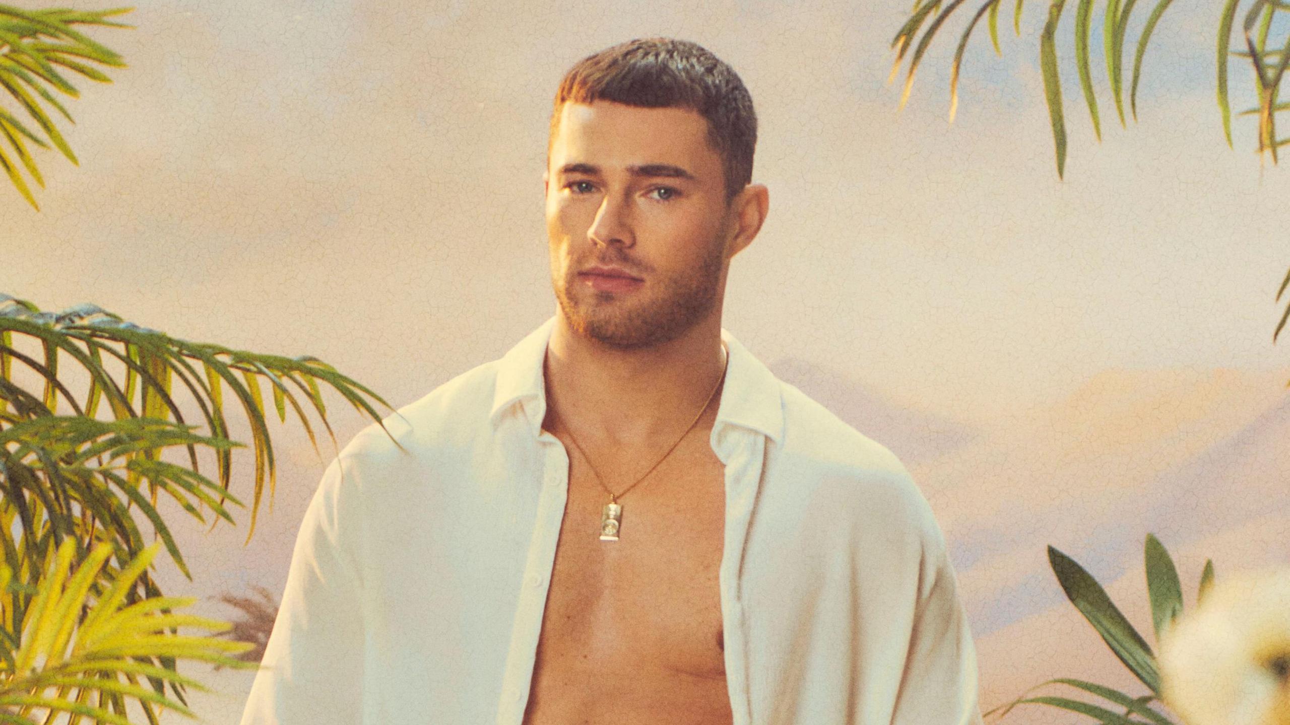 Curtis Pritchard in a promo shot for the new series of Love Island All Stars. Curtis, who has cropped brown hair, wears an unbuttoned white shirt and gold necklace. He's pictured against a dusky sky surrounded by green foliage. 