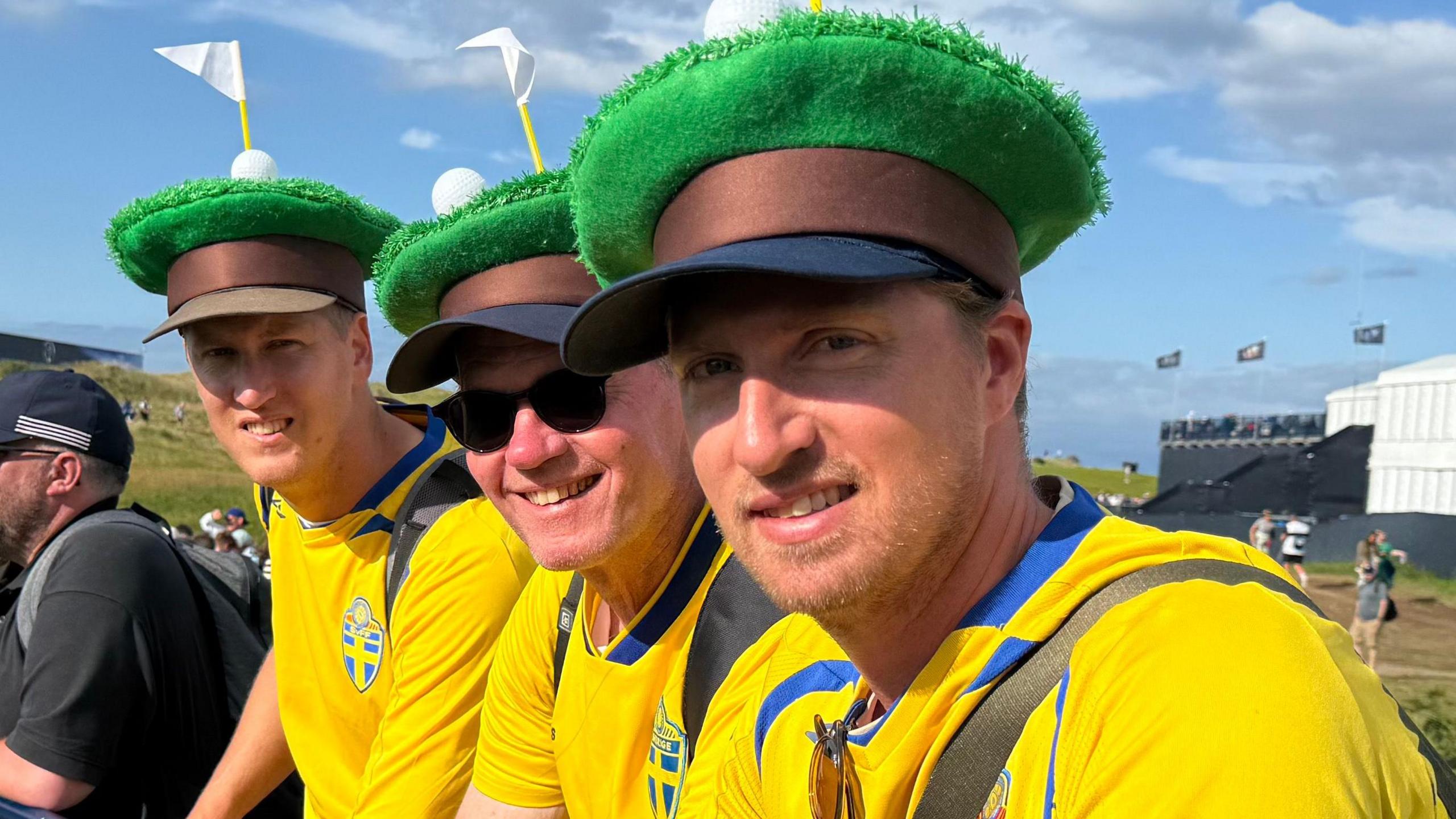 Three men are standing sideways shoulder to shoulder and looking sideways into the camera. They are all smiling and wearing green hats that look like fake grass with a brown band below and a navy visor. They are all dressed in yellow t-shirts with a blue trim and a crest of the Swedish flag on the top front left hand side of their t shirts