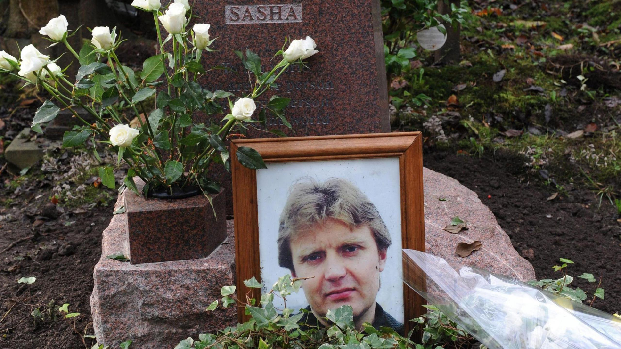 Alexander Litvinenko's grave. A framed photograph of him is in front of the headstone and there is a vase of white roses. The headstone has a plaque reading "Sasha" on it - a nickname for him.