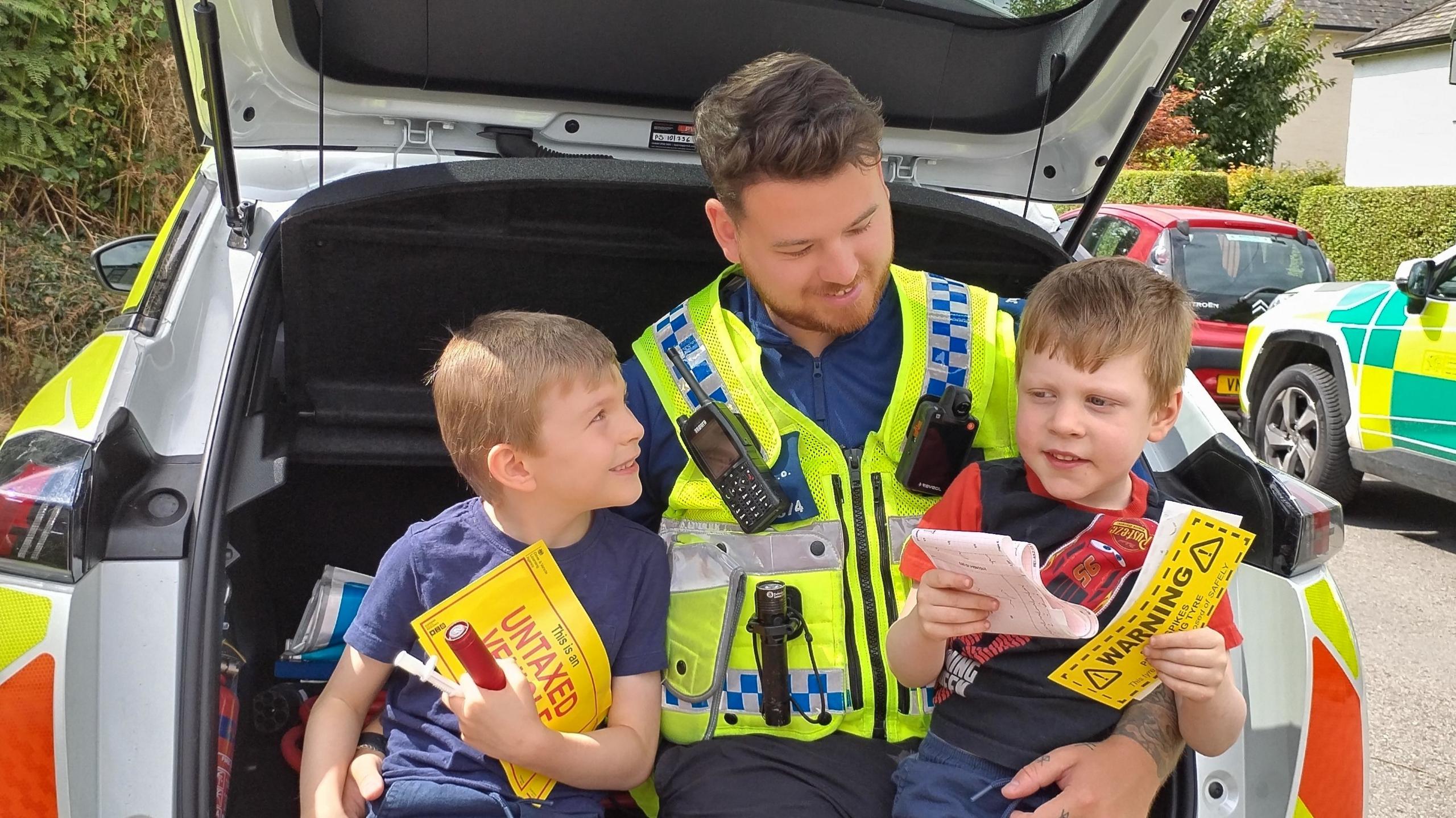 Joshua Carey in his community support officer uniform sat in the boot of a police vehicle with his arms round Macsen and Trystan on either side of him