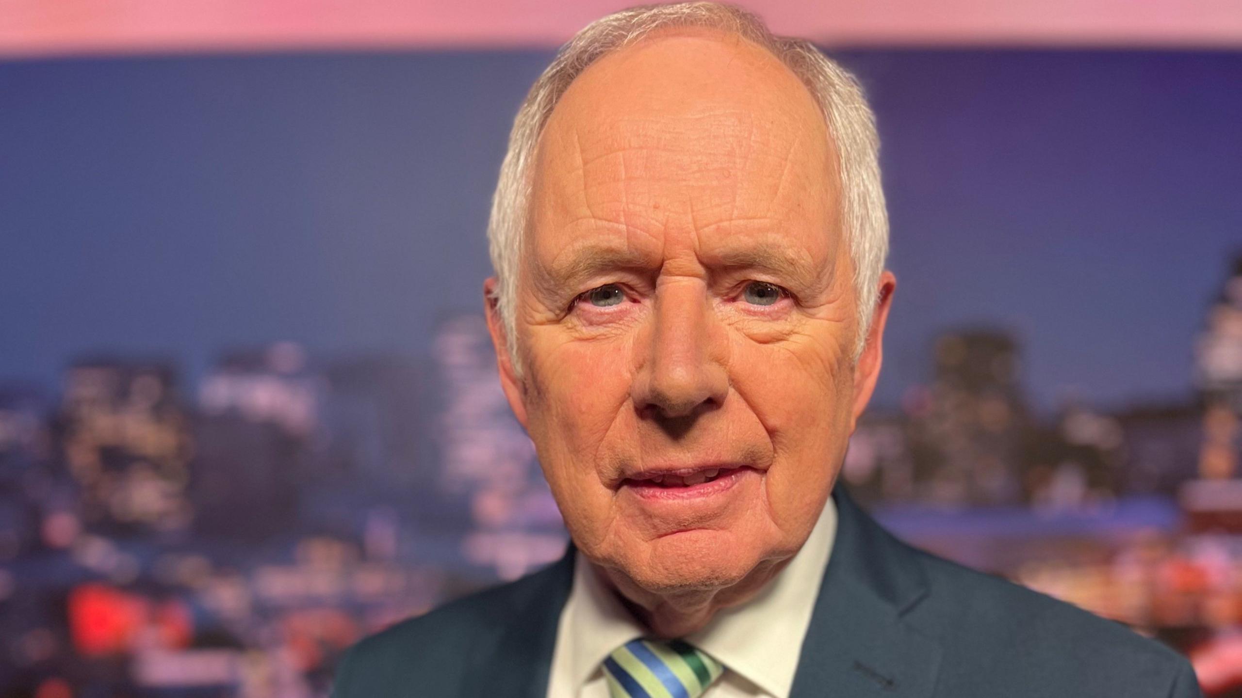 Reporter Nick Owen. He is balding with white hair at the sides and has blue eyes. He is wearing a blue suit jacket with a white shirt, and blue and green striped tie. He is looking at the camera and behind him is a blurred city skyline. 