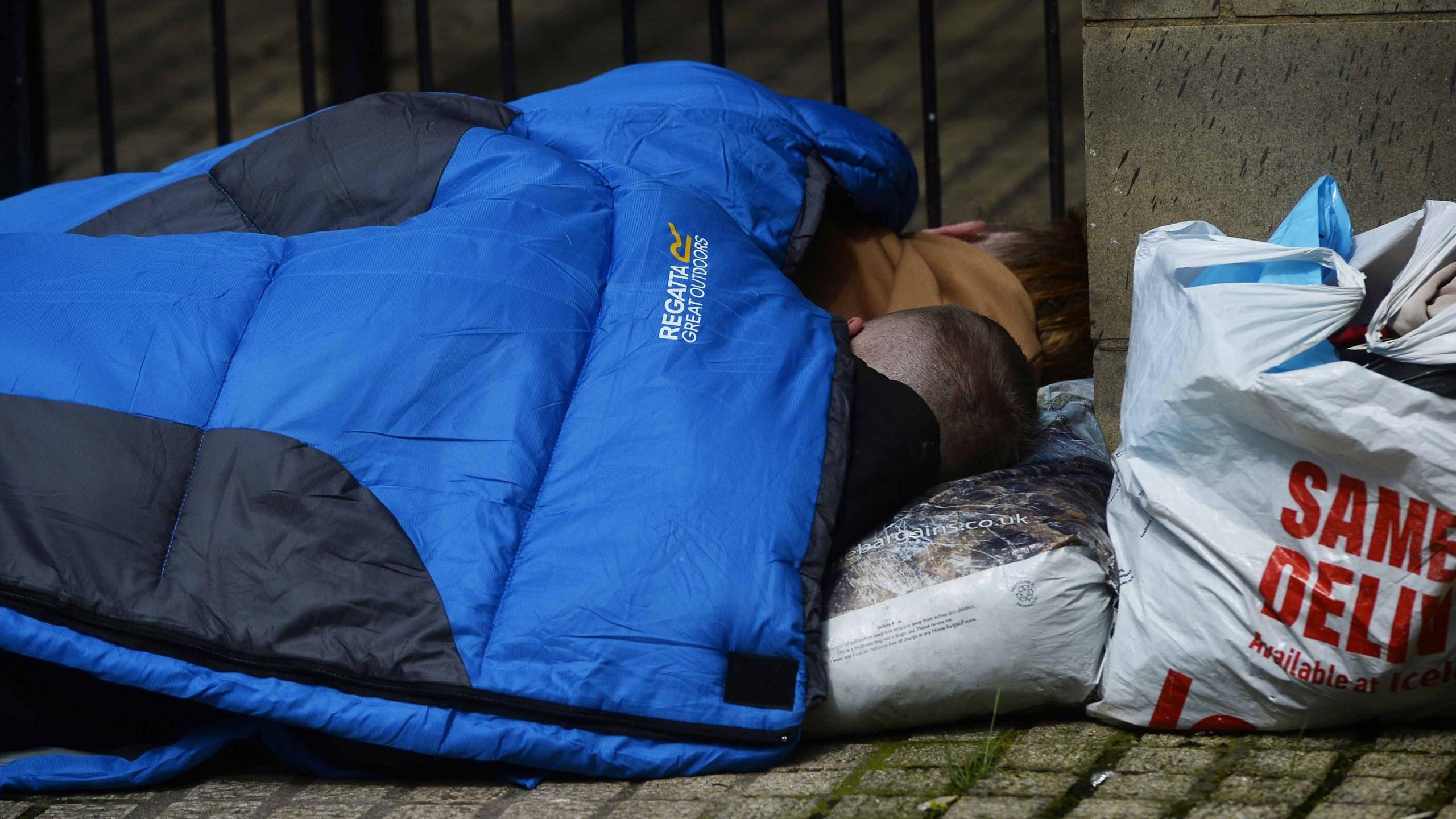 Rough sleeper under a blue sleeping bag resting their head on a plastic bag