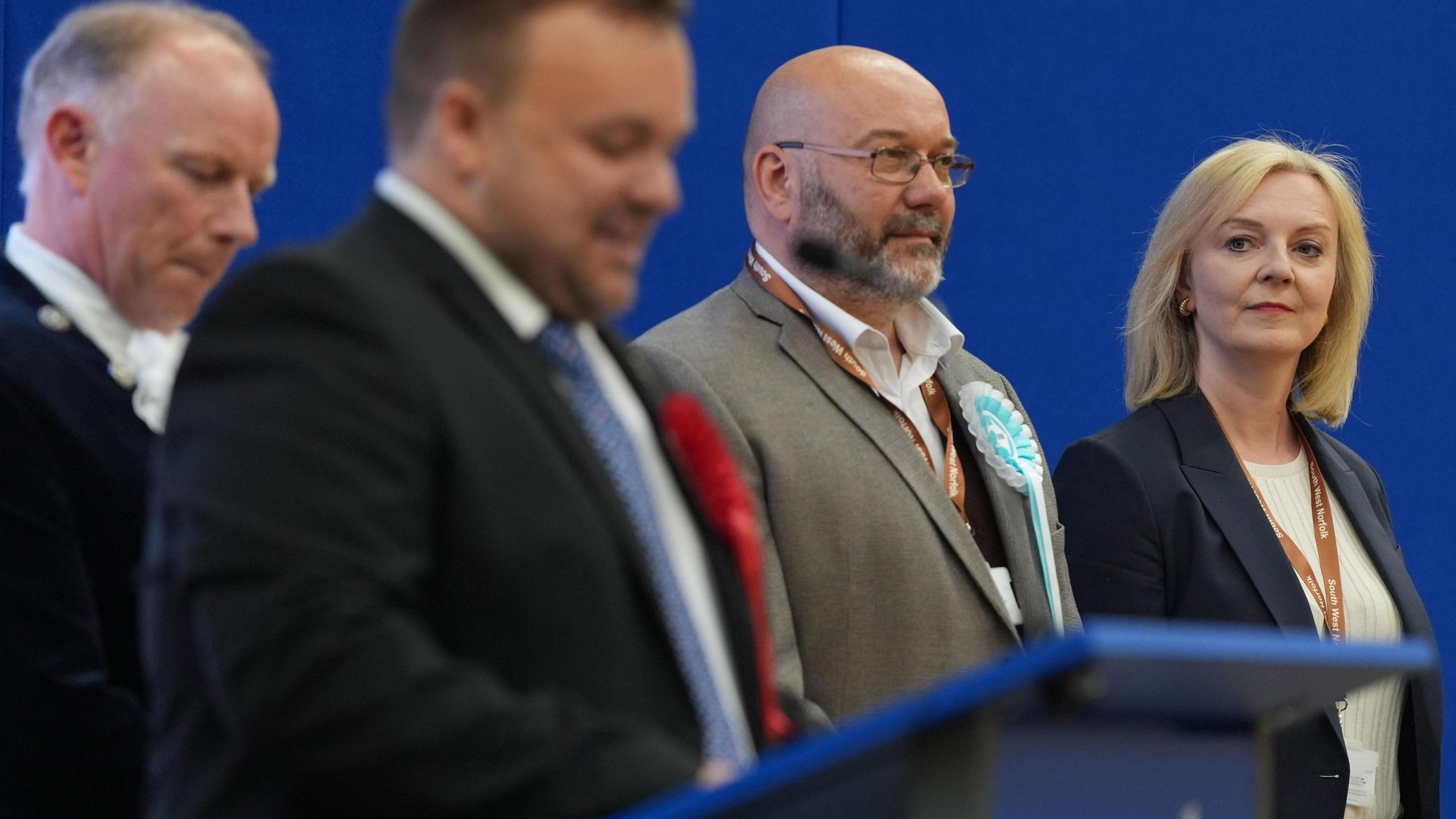 Two men in suits look on as Terry Jermy delivers a speech from a podium, with Liz Truss standing to the right and looking on too.