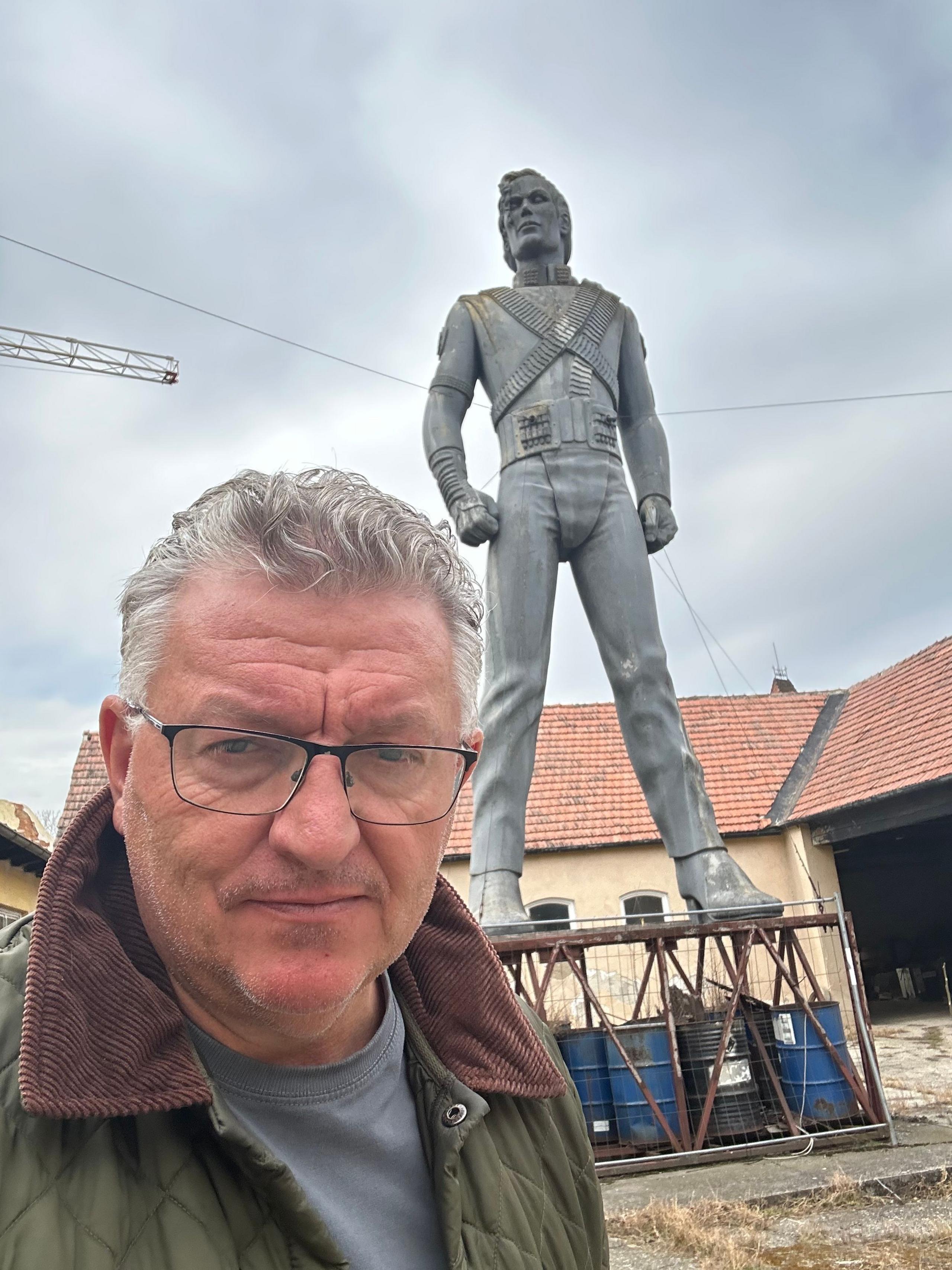 A man with glasses and silver hair is taking a selfie in front of a Michael Jackson statue perched on a base made of oil drums. Parts of the statue look brown and stained white.