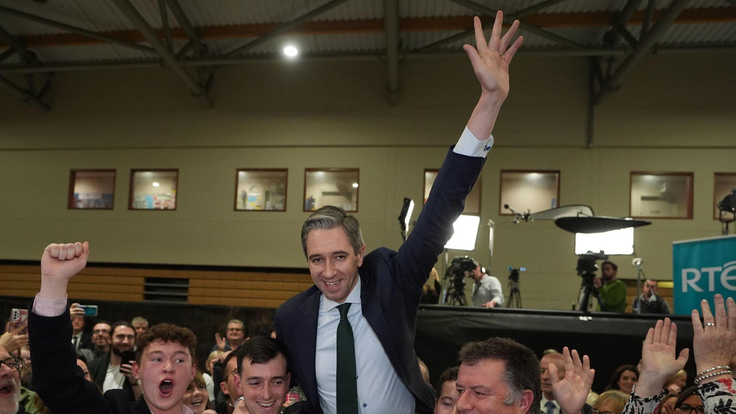 Simon Harris celebrates at a count centre. He is wearing a blue jacket and blue tie and white shirt. 