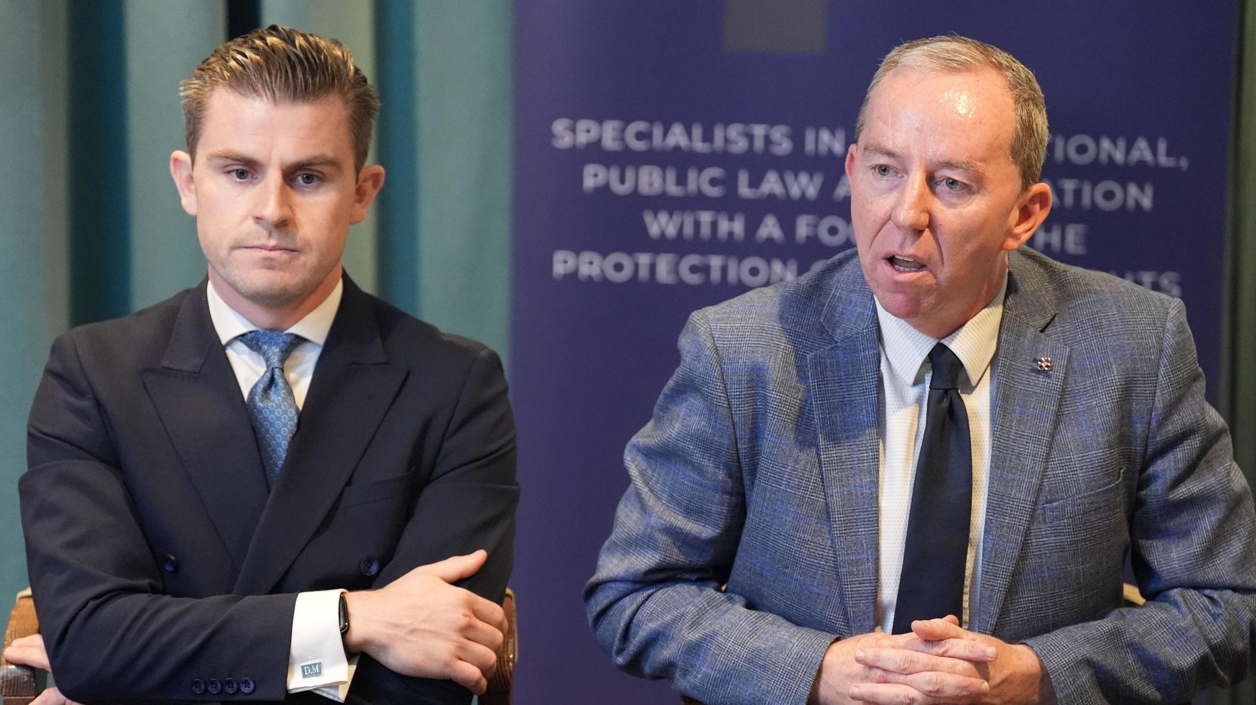 Peter Corrigan and Darragh Mackin speaking at a news conference. Peter is wearing a grey suit and Darragh is wearing a black suit