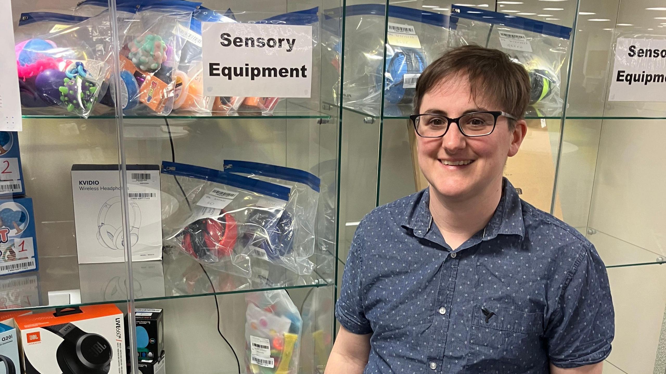 A person in a blue short-sleeved shirt stands smiling in front of a glass cabinet filled with noise cancelling headphones and fidget toys. A sign reading 'Sensory Equipment' is taped to the front of the cabinet.