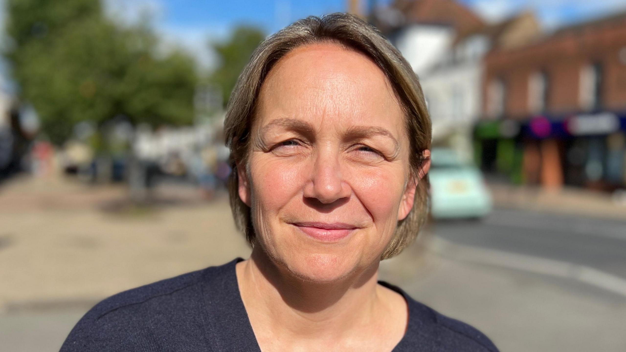 Kerry is wearing a dark T-shirt on a quiet high street scene. She has short blonde hair.