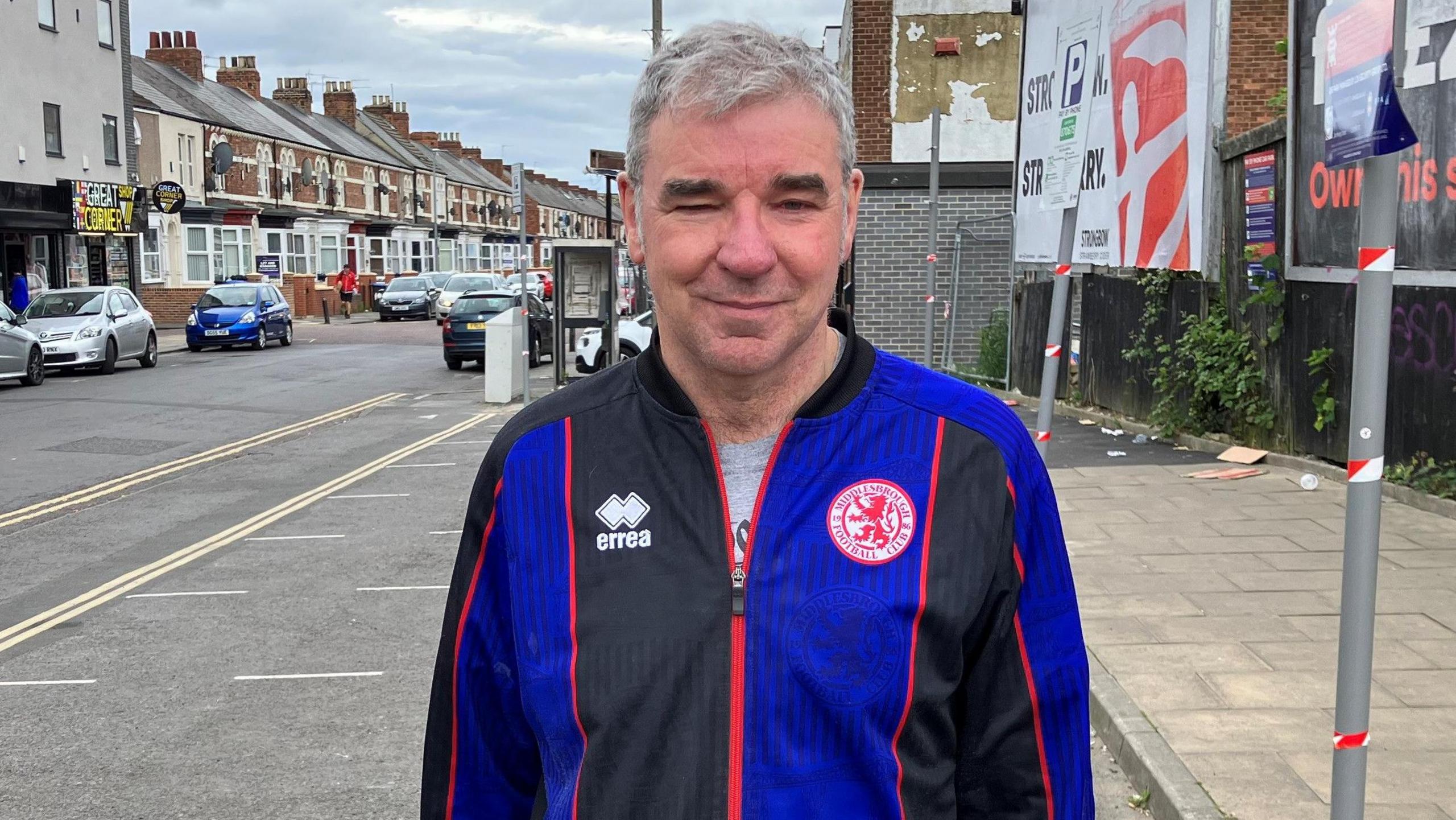 Robert Nichols standing on Linthorpe Road. He has short grey hair, brushed to his right, and is wearing a blue-and-black Middlesbrough FC tracksuit top. A row of terraced houses can be seen in the background on the left, while on the right are two advertising billboards.