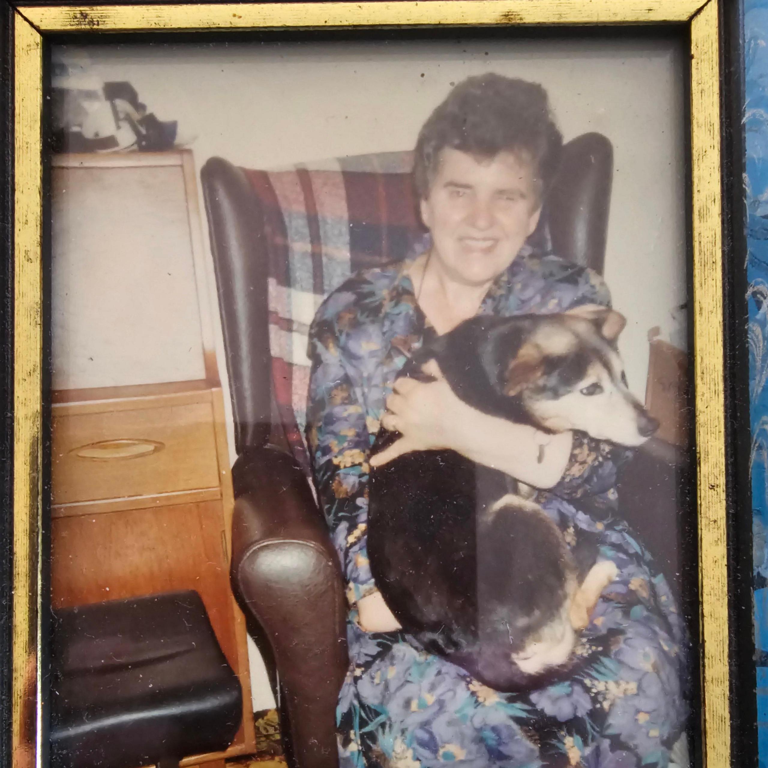 A photo in a gold frame of a middle-aged woman with short black hair grinning at the camera as she holds a black and brown dog in her arms. She sits in a brown leather armchair and is wearing a black and blue floral dress.