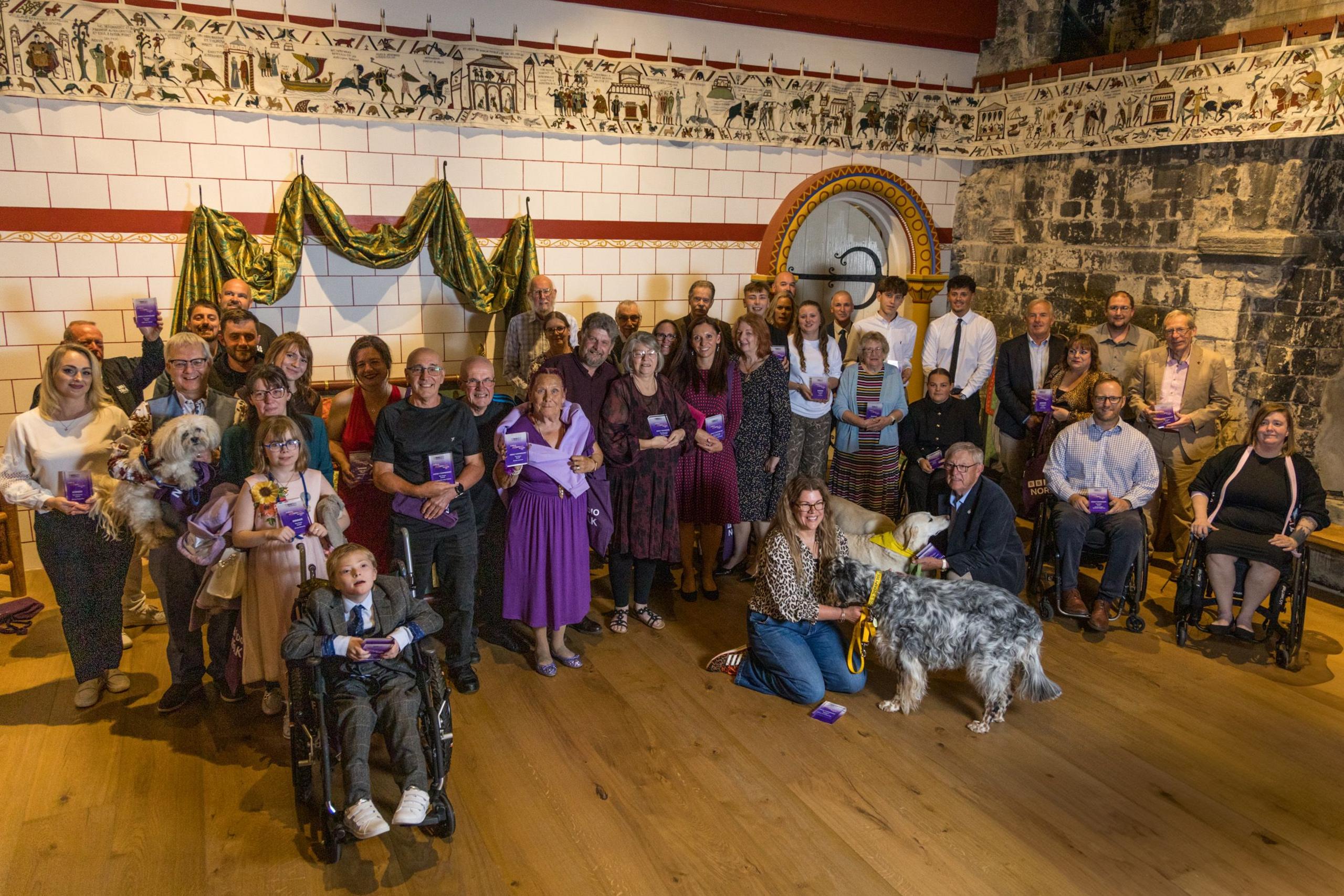 A group of dozens of people stand and sit in front of a white brick wall. Many of them are holding glass awards.
