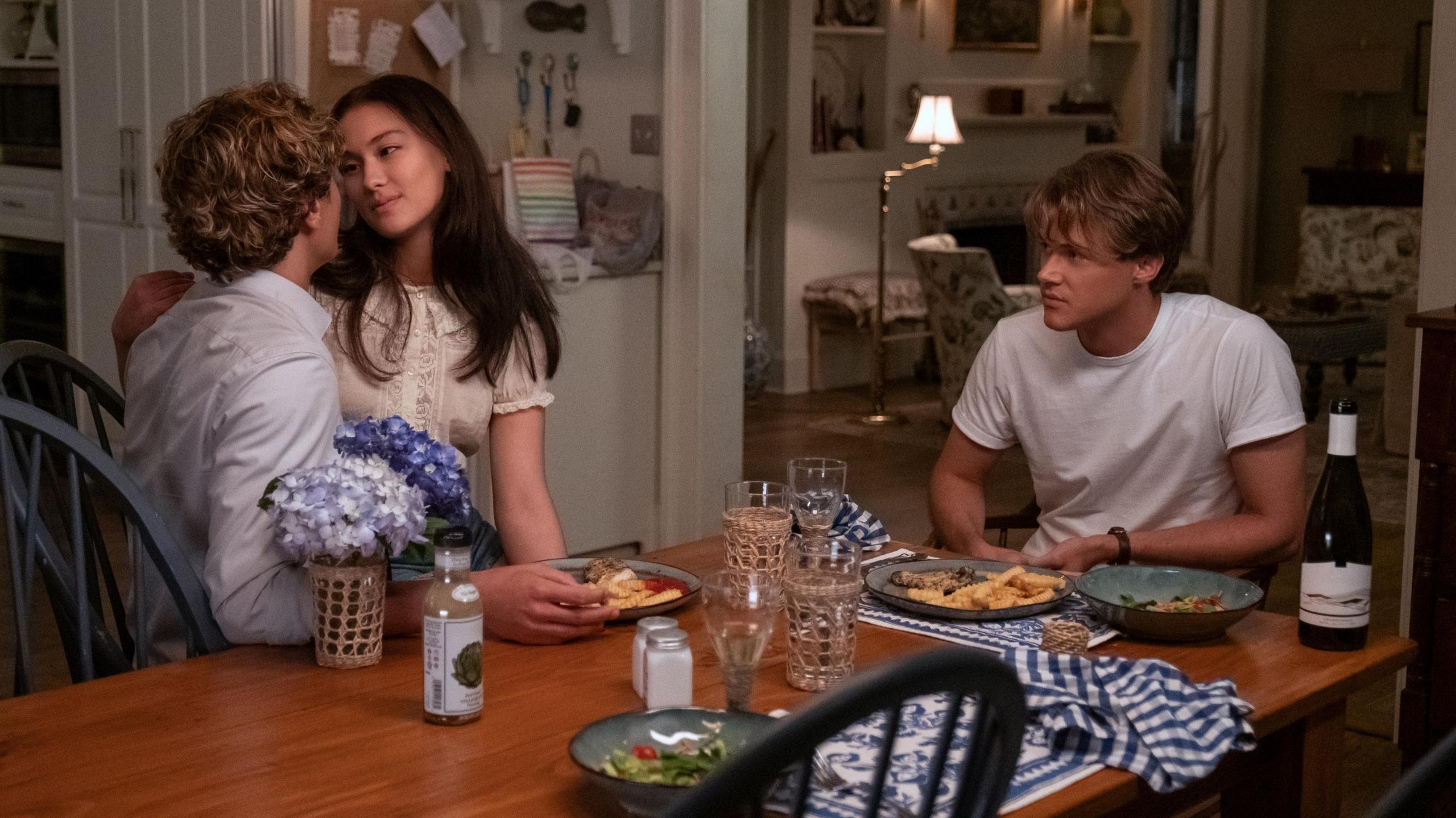 Belly sitting on Jeremiah's lap and gazing lovingly into his eyes at a dining table, with Conrad looking on jealously