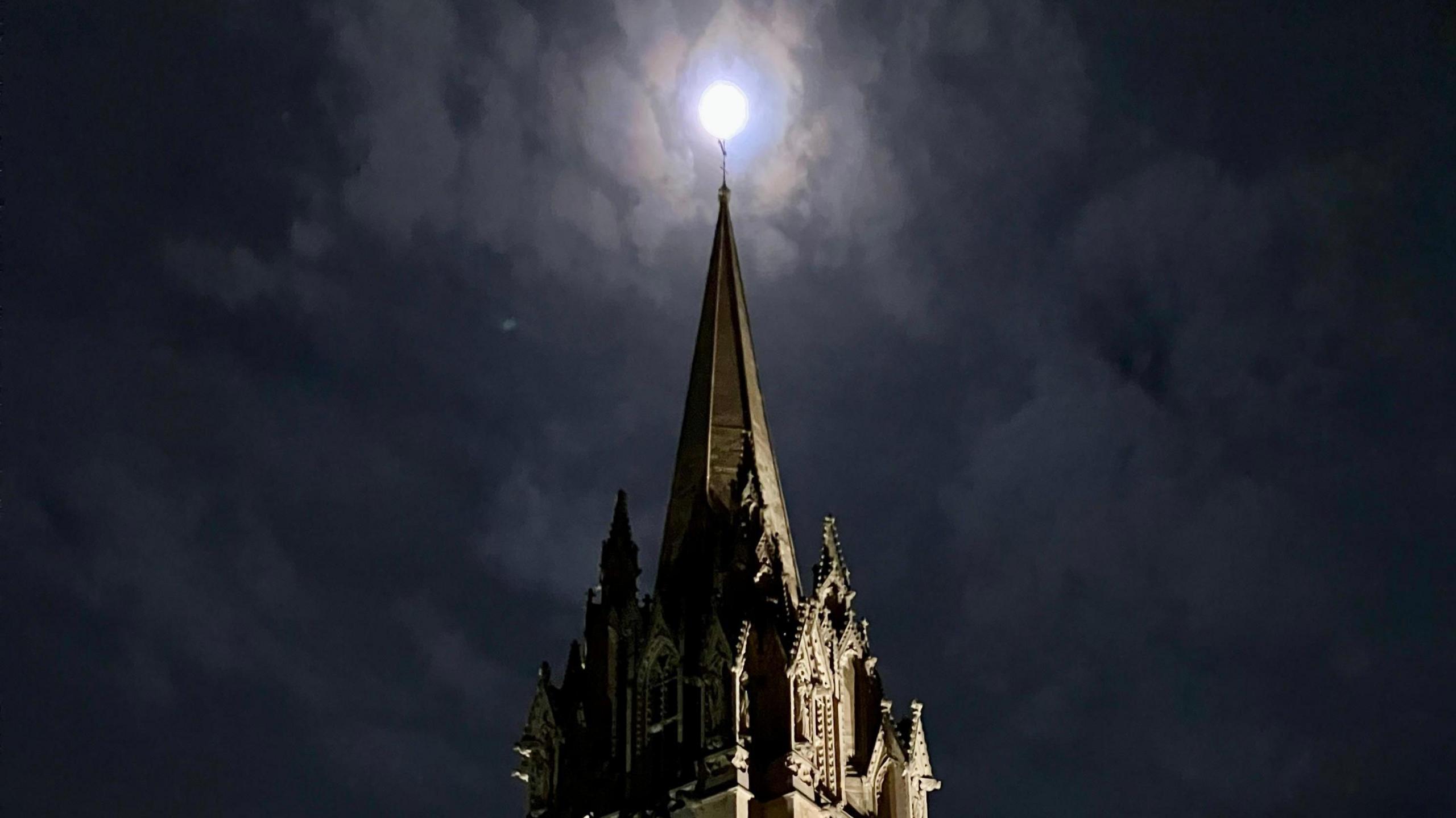 The spire of a building against a night sky with a bright white moon appearing at its tip.