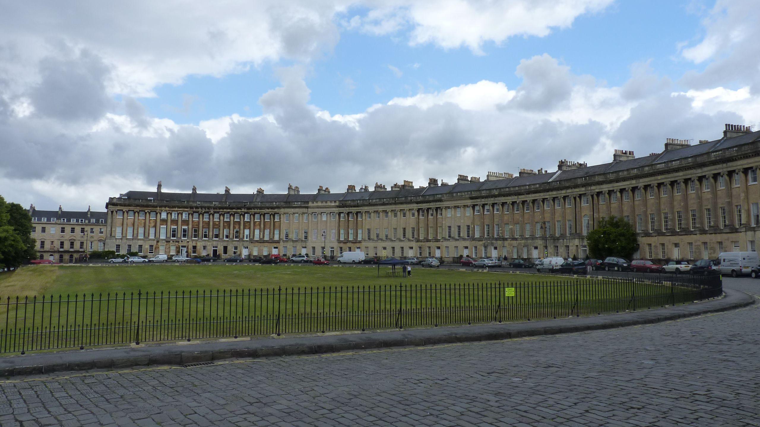 A side view of Royal Crescent showing homes in a semi-circle, taken from the road. There is a green lawn and cars are parked outside of the row of homes.