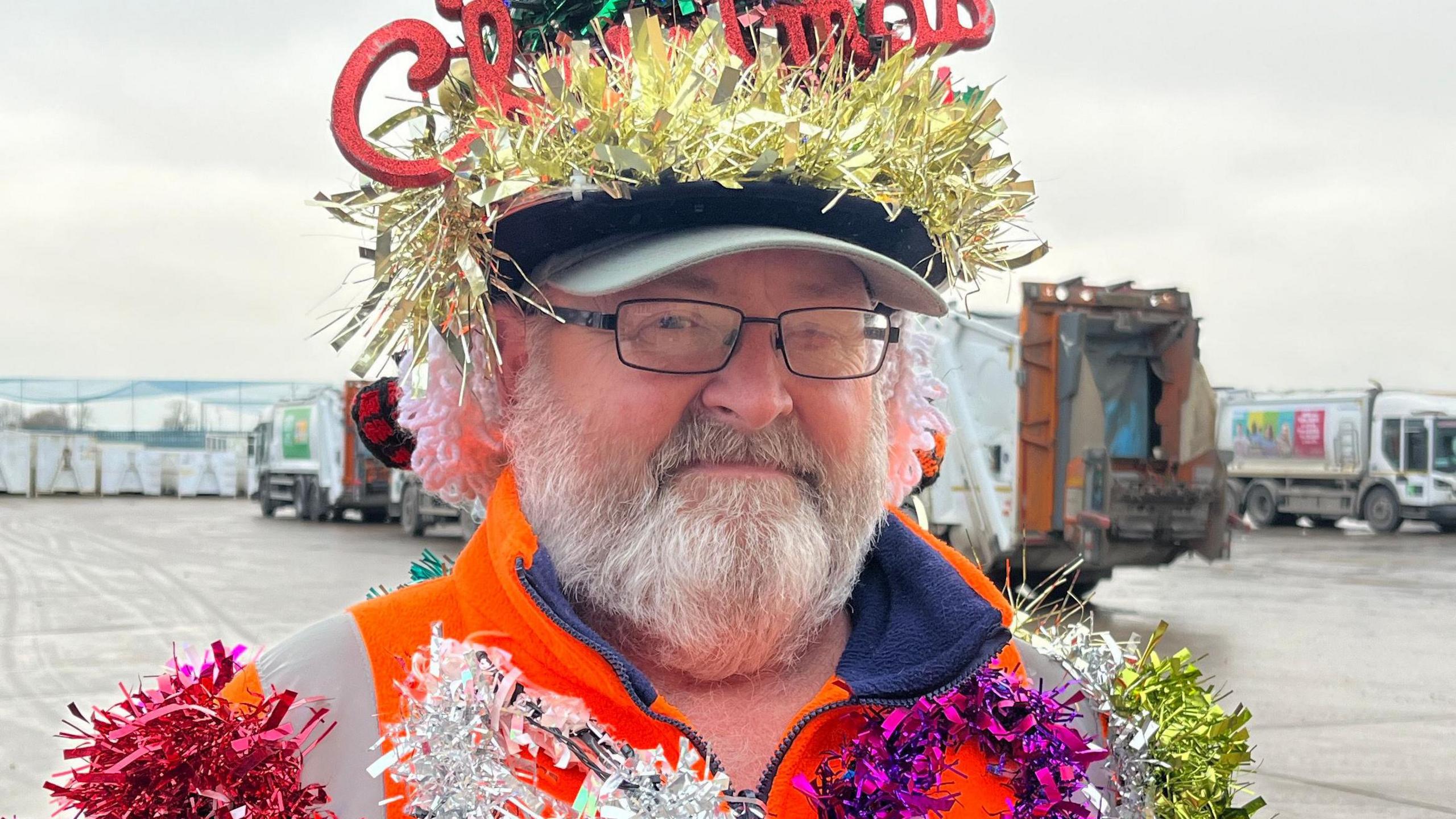 Close up of Lenny Edward's face. He has a white beard, black thin framed glasses and is stood at a recycling centre in front of bin lorries. On his head is a  baseball cap covered in gold tinsel with the words Merry Christmas sticking up from the top of his hat in red glittery writing. Over his high visibility orange uniform  he has pink, white, purple, green and gold tinsel.