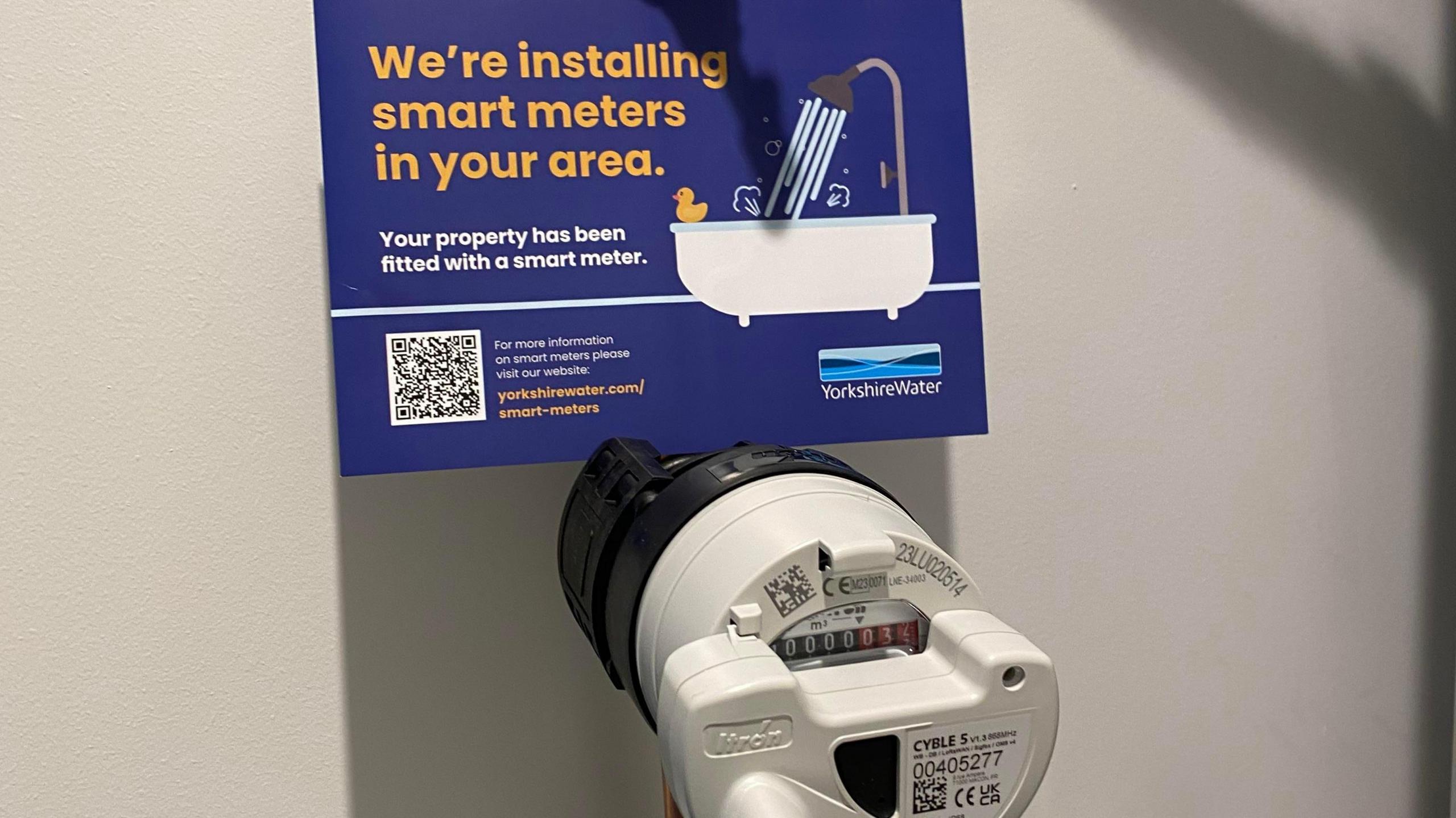 A smart water meter installed on an internal copper stop tap, it is a white plastic circular device with a meter reading on it. There is a Yorkshire Water card above which reads, we are installing smart meters in your area.