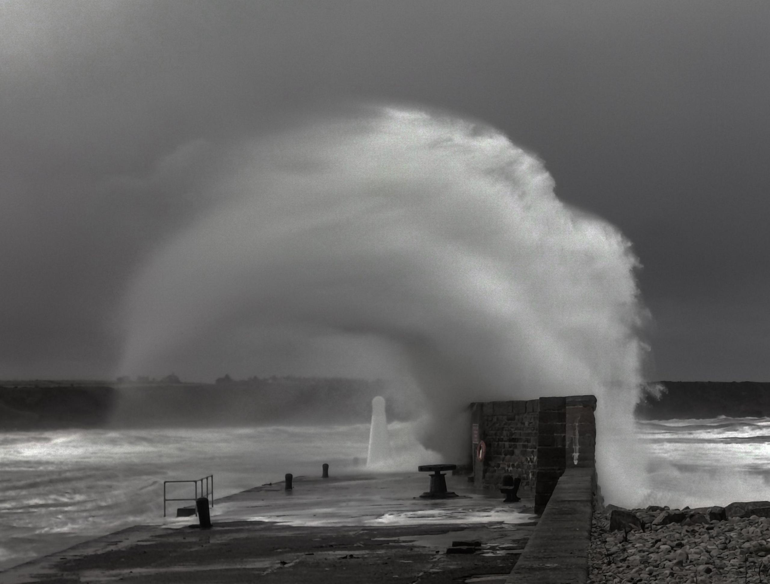 A black and white photograph of a large wave crashing against a sea wall