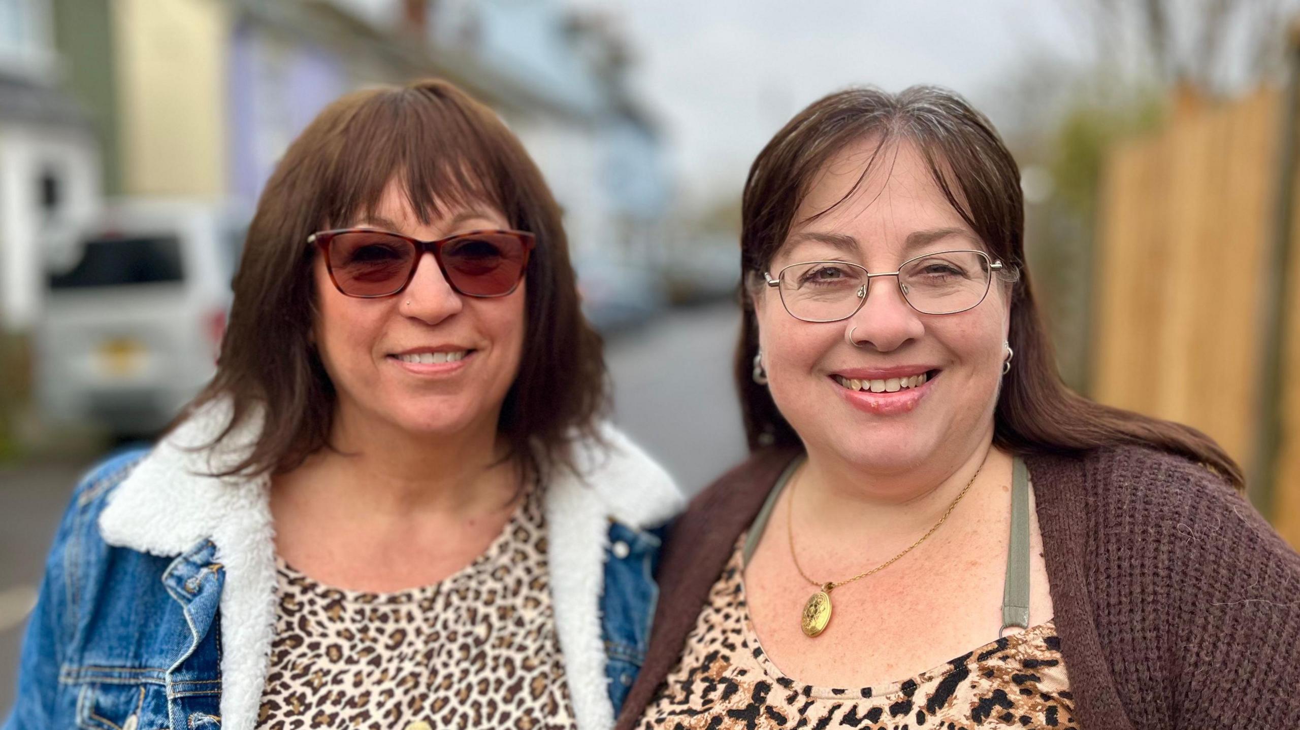 Two women smiling straight at the camera. One is wearing sunglasses, a leopard print top, and denim jacket. The other is wearing glasses, a leopard print top, and a brown cardigan.