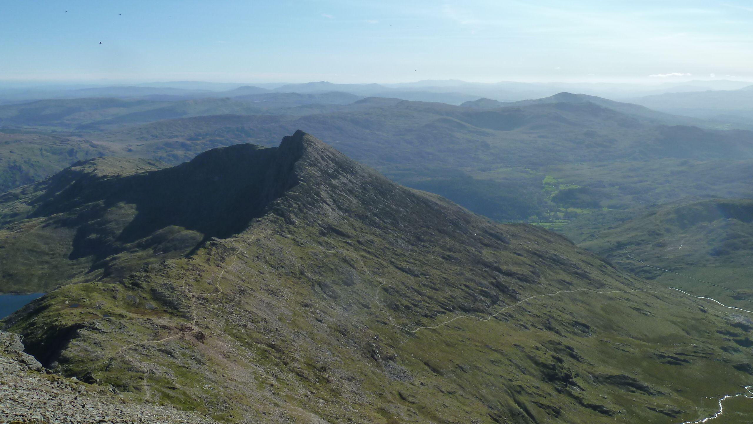 A picture of the Eryri (Snowdonia) national park from Yr Wyddfa (Snowdon). There are several mountains on the horizon with a mountain in the foreground. Most of the land is green with some rocky outcrops and a few paths visible