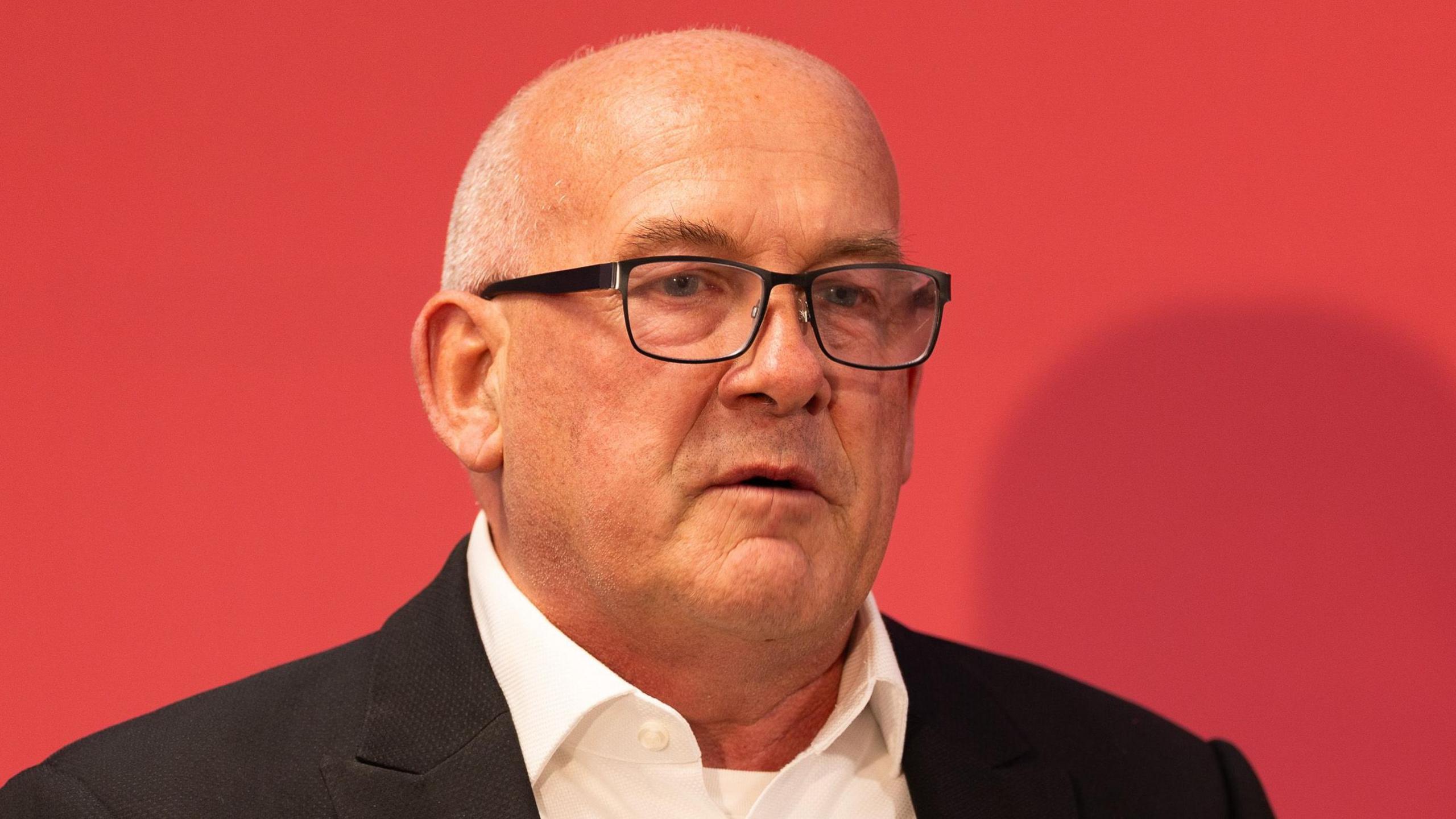 A close-up of Richard Collier-Keywood in dark jacket and open white shirt in front of a red background