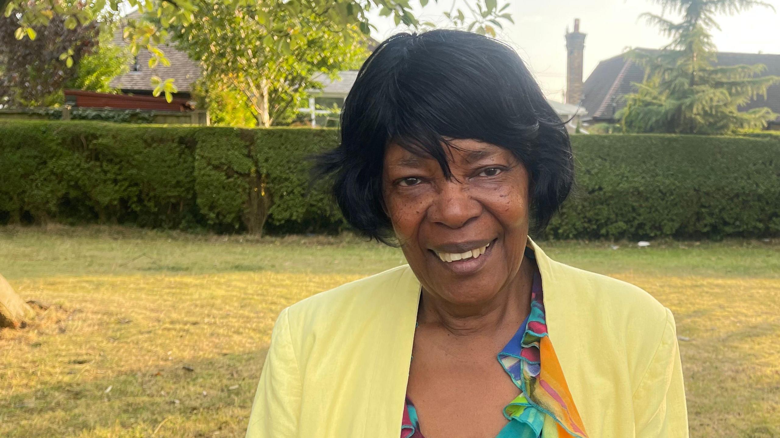 A woman with black hair, a yellow blazer and a bright, colourful blouse is stood in a park. Behind her is a hedge, a tree and two houses.