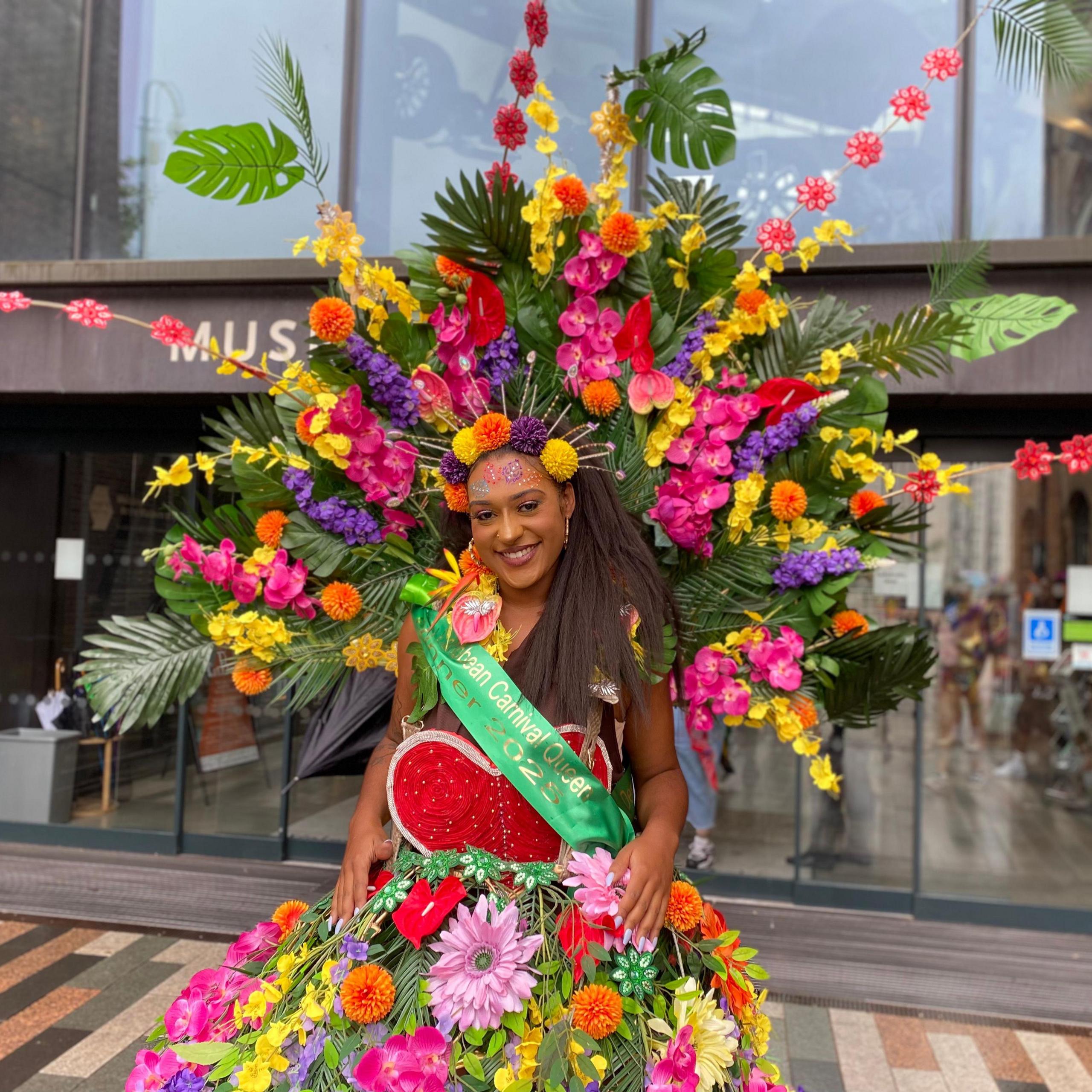 A woman in a huge floral dress with palm fronts and huge flowers attached, and a green sash