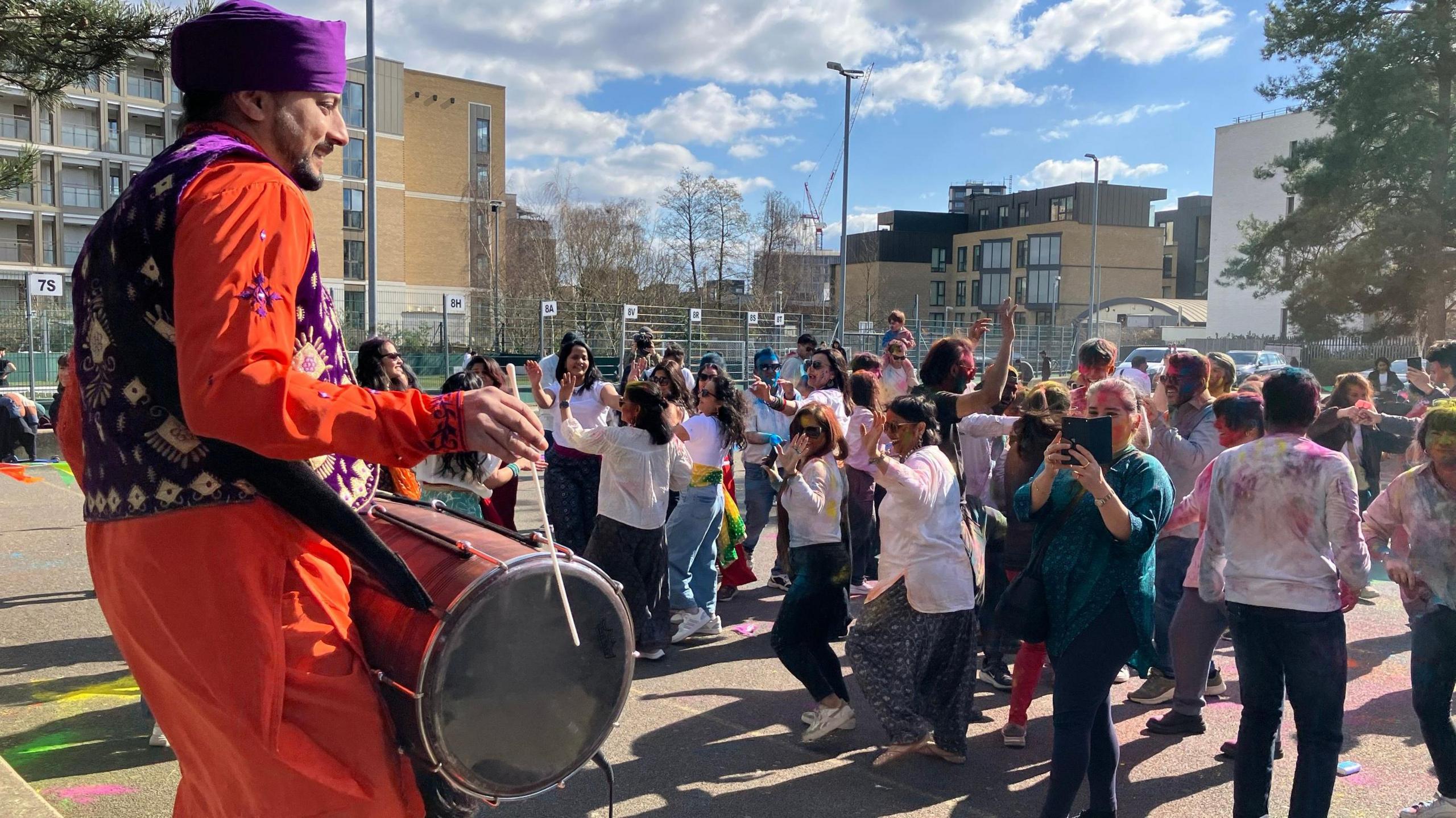 A drummer dressed in bright orange plays his instrument as a group of young people dance. 