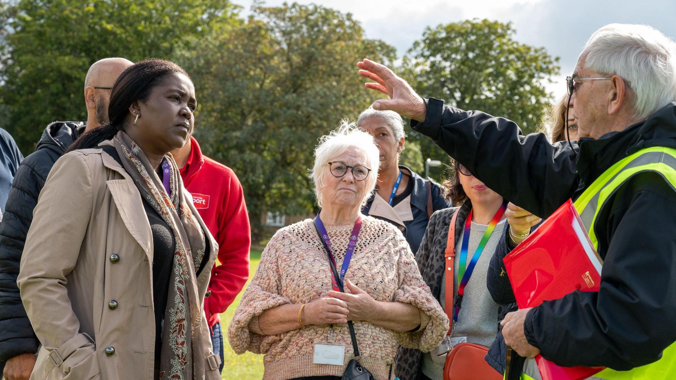 A group of people gather in a park listening to a man in a high visibility jacket who is pointing off to his right. 