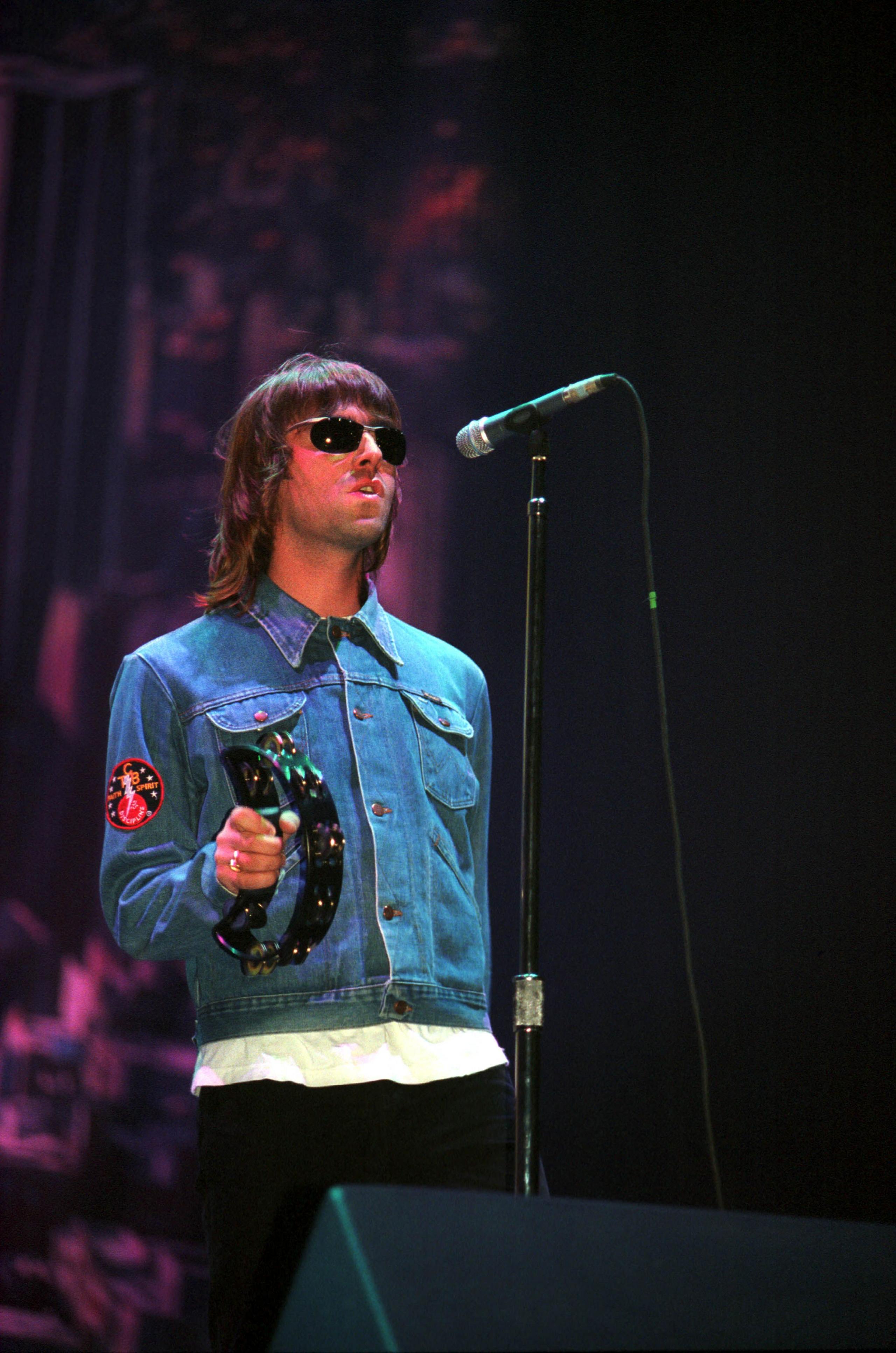 Liam Gallagher performing at Glasgow Green, on stage wearing a blue denim jacket holding a tamborine