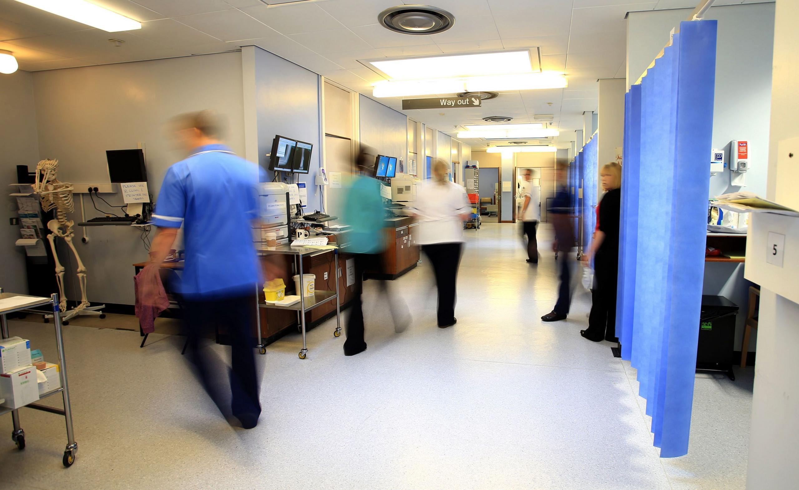 A busy hospital corridor with medical staff in motion, creating a blurred effect. Staff members in blue uniforms can be seen walking, with medical equipment and computer screens visible along the walls. The corridor has fluorescent lighting and a clinical appearance with white walls and blue accents.