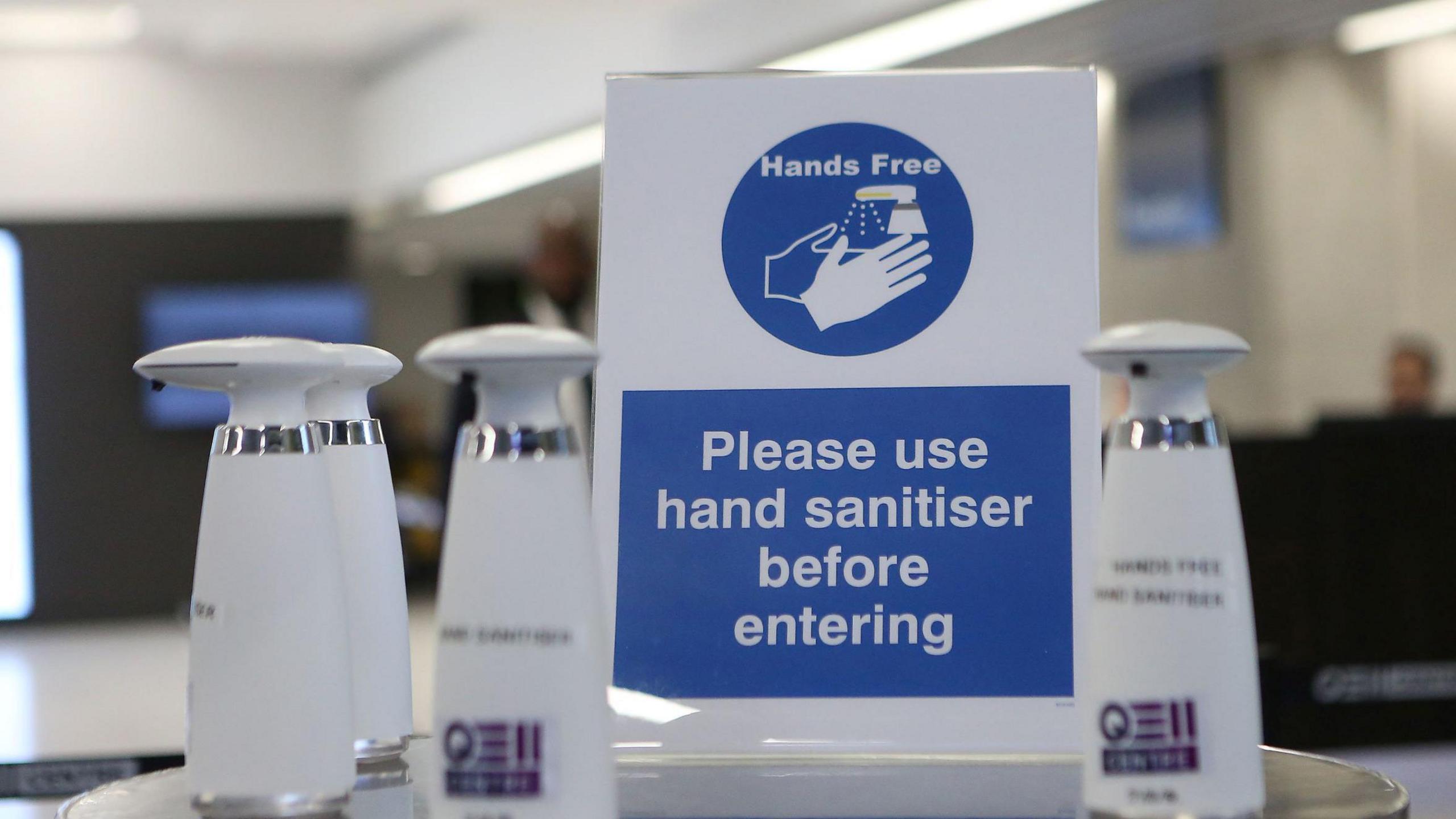 A table with hand sanitiser products sitting on it. A sign reads Hands Free, with a picture of the sanitiser spraying onto hands without being touched, and text reading PLEASE USE HAND SANITISER BEFORE ENTERING.