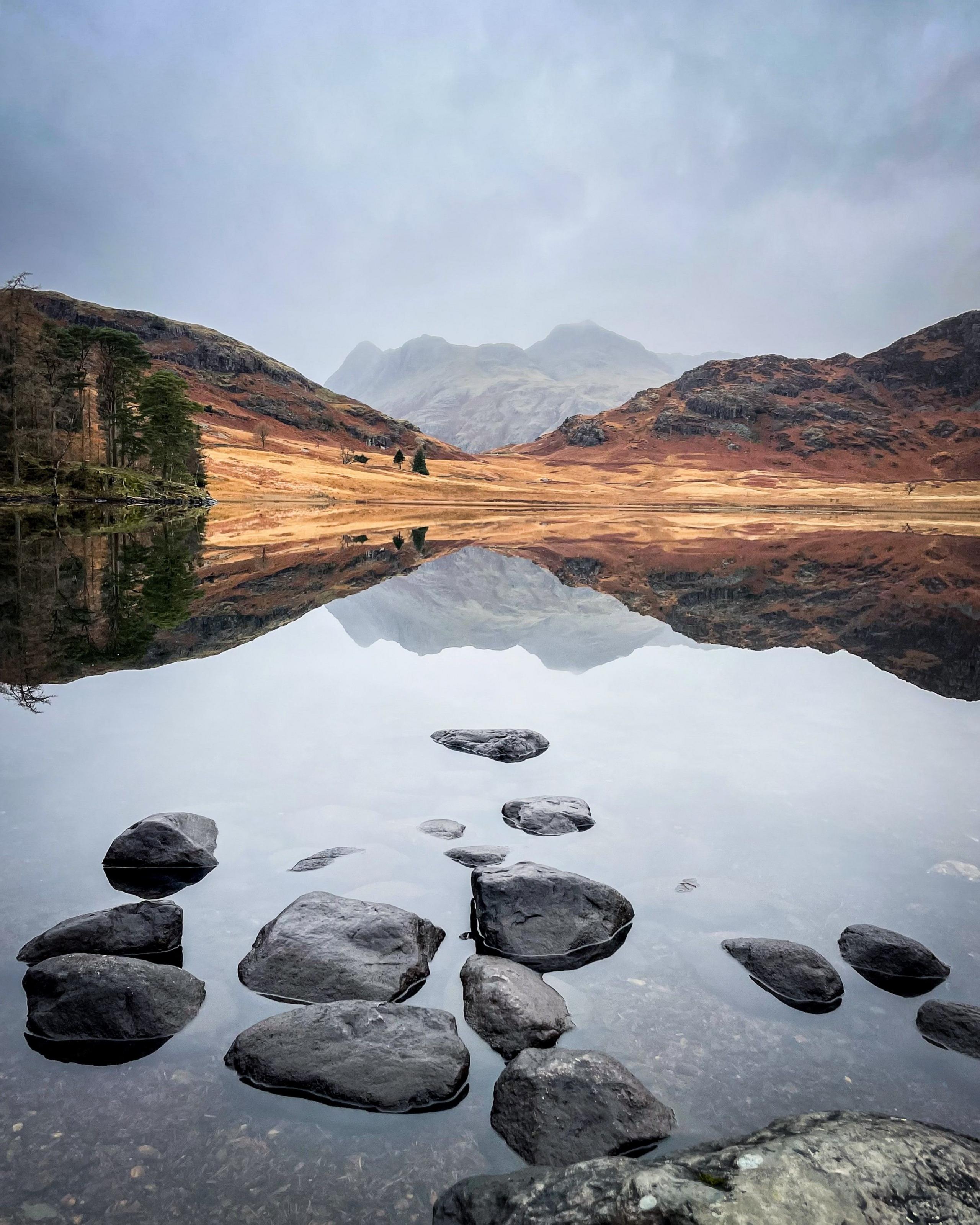 A perfectly still tarn, with a yellow and green hill beyond and distant craggy mountain reflected perfectly.