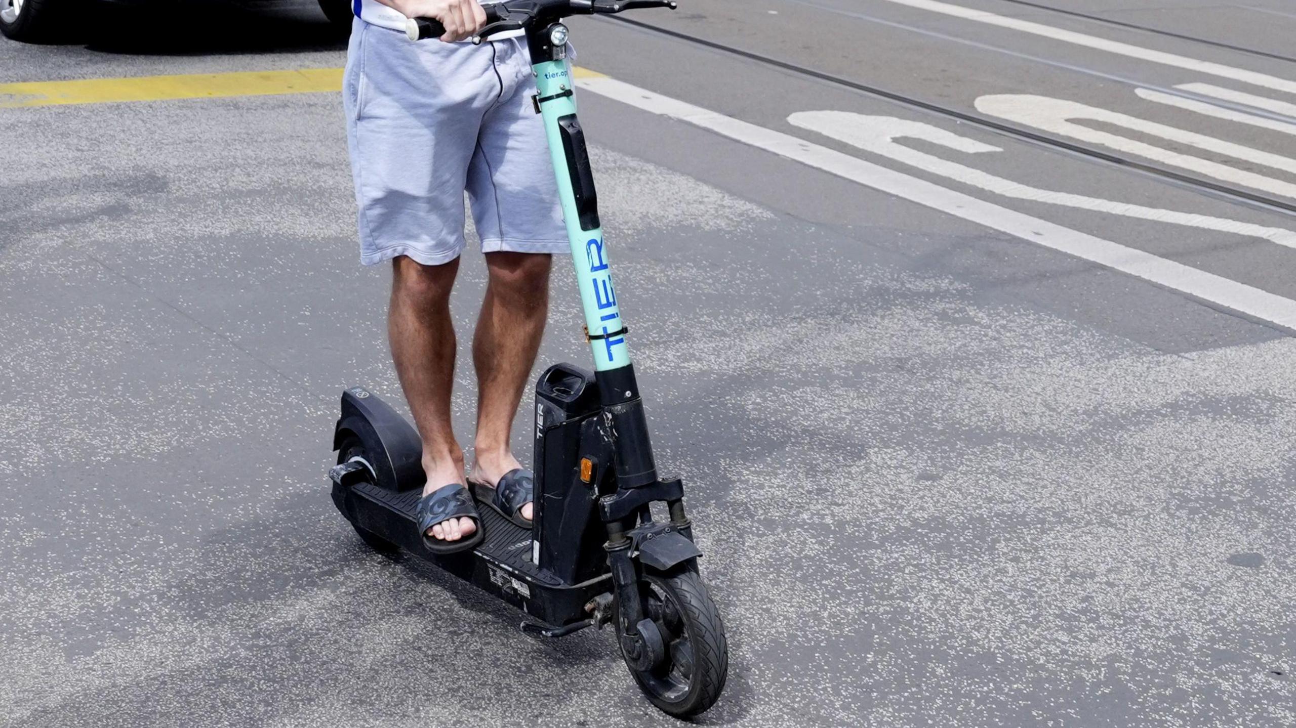 The bottom half of a male rider on an adult electric scooter.  He is wearing shorts and flip flops.