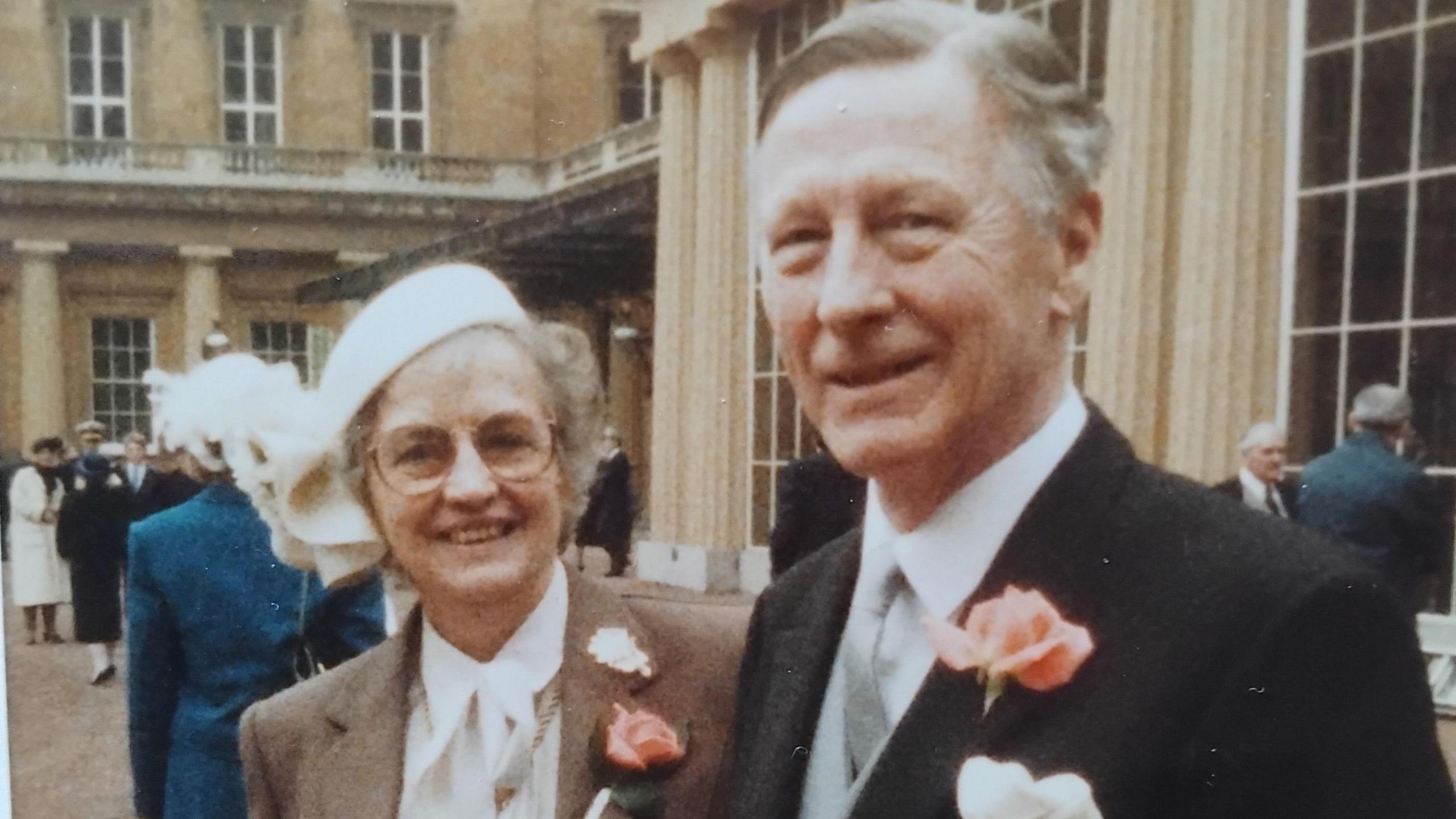 Jack and his wife Betty stand outside Buckingham Palace smiling at the camera, with other people dotted around behind them. He wears a black suit jacket with a salmon coloured rose in the lapel, and a white shirt with silver tie. 