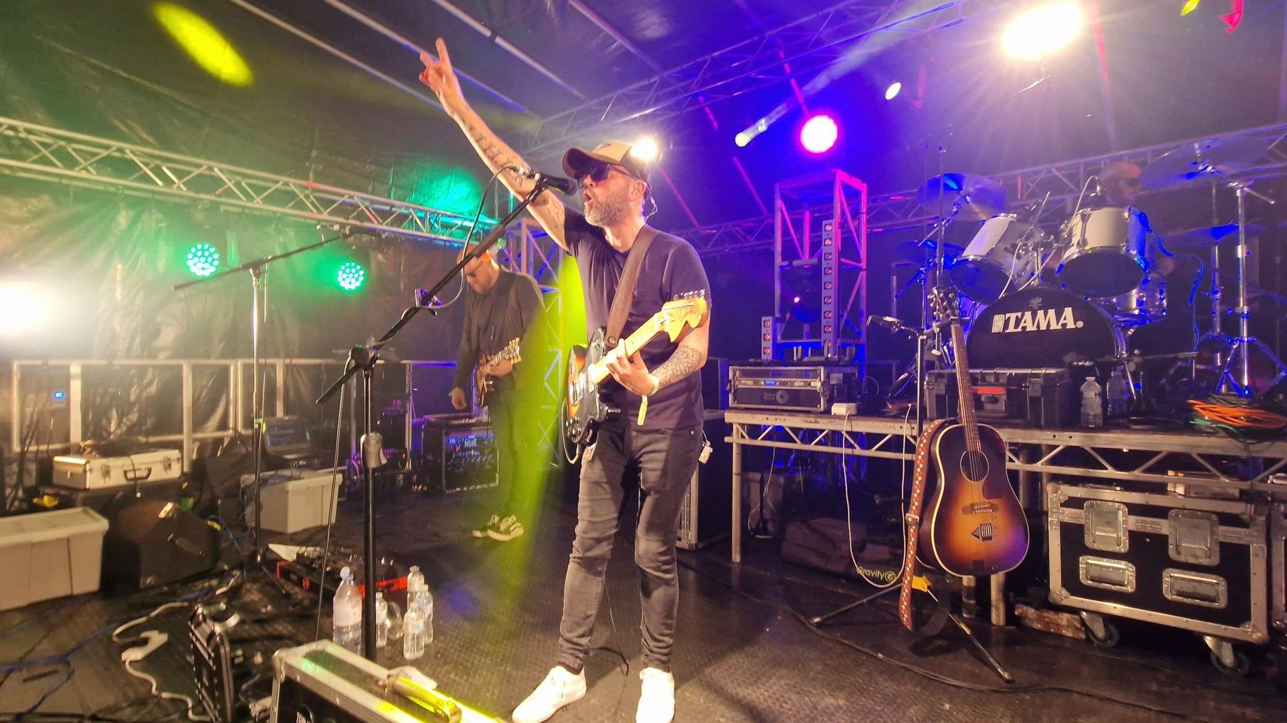 A man who is wearing a black t-shirt and jeans is standing on a music stage. He is playing a guitar and singing into a microphone. Multi-coloured flashing lights surround him.