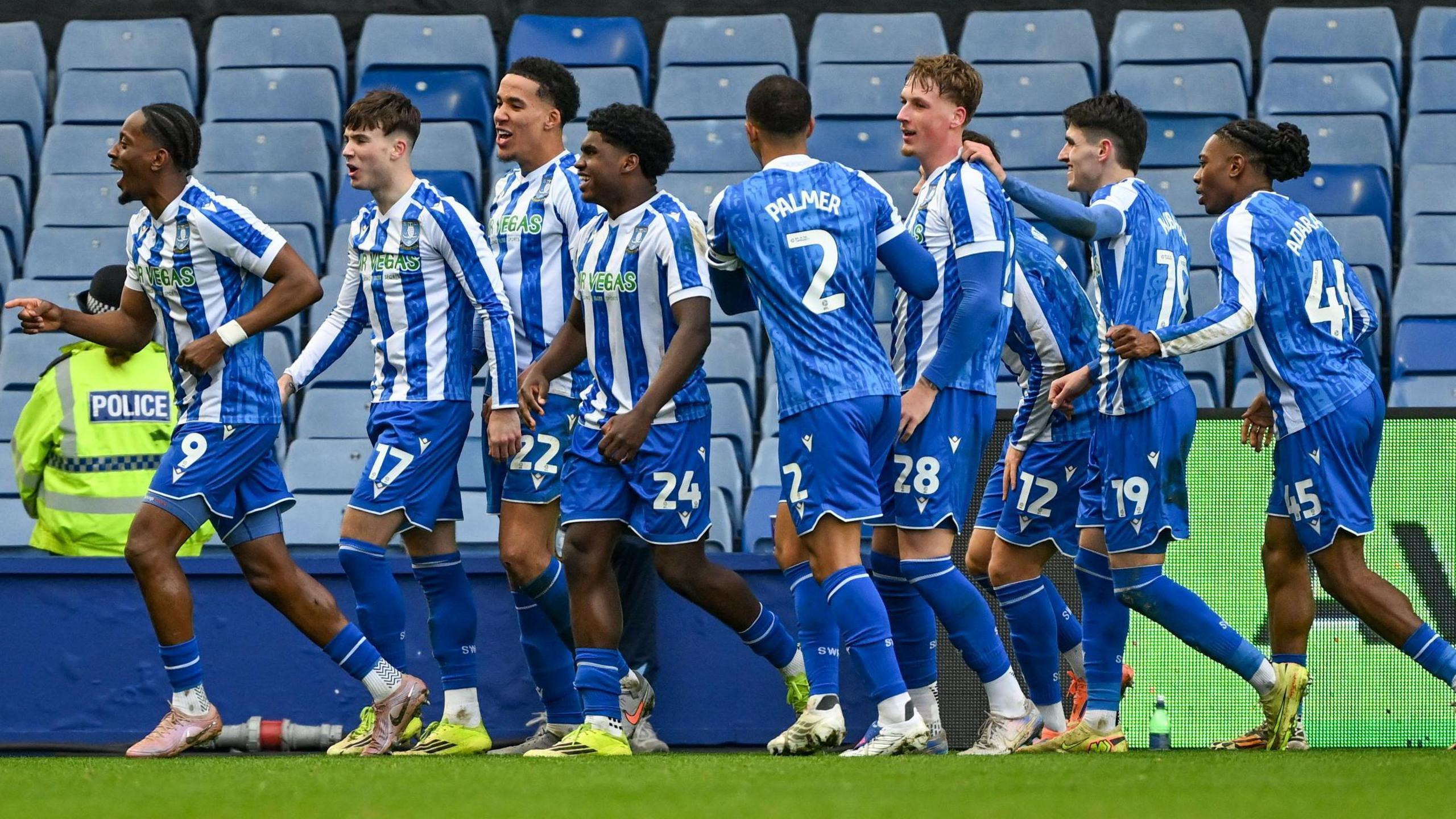 Nine Sheffield Wednesday players, wearing their home strip of blue and white vertical striped shirts, blue shorts and socks, celebrate scoring against Millwall in front of empty blue seats, with a police officer sitting on the perimeter wall