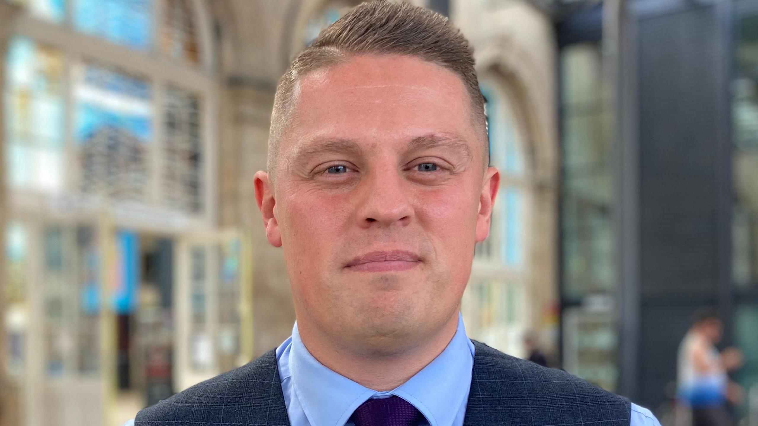 Adam Shone wearing a dark blue waistcoat and tie over a blue shirt