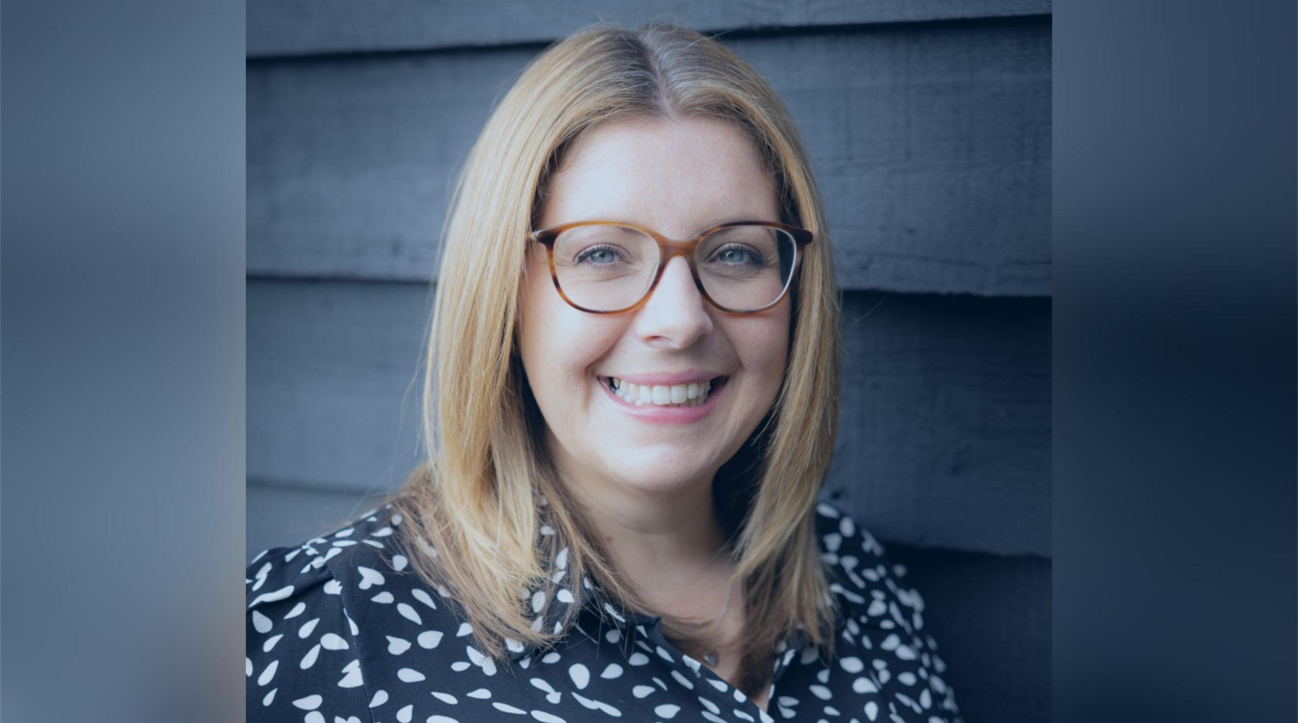 Hannah Bloom with long blonde hair and glasses, wearing a black dress with white dots, smiling at the camera.