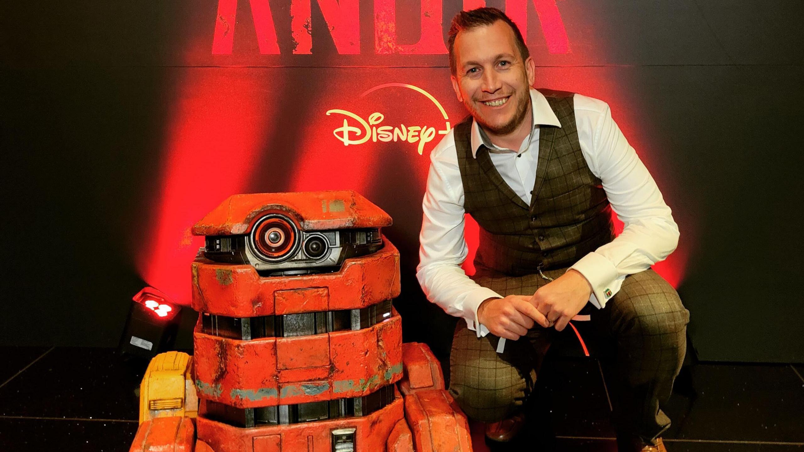 Chris Saunders crouches next to an replica Star Wars droid. The droid is about two feet tall, with a distressed orange paint look, it has a single eye-like camera towards to top of the body. 