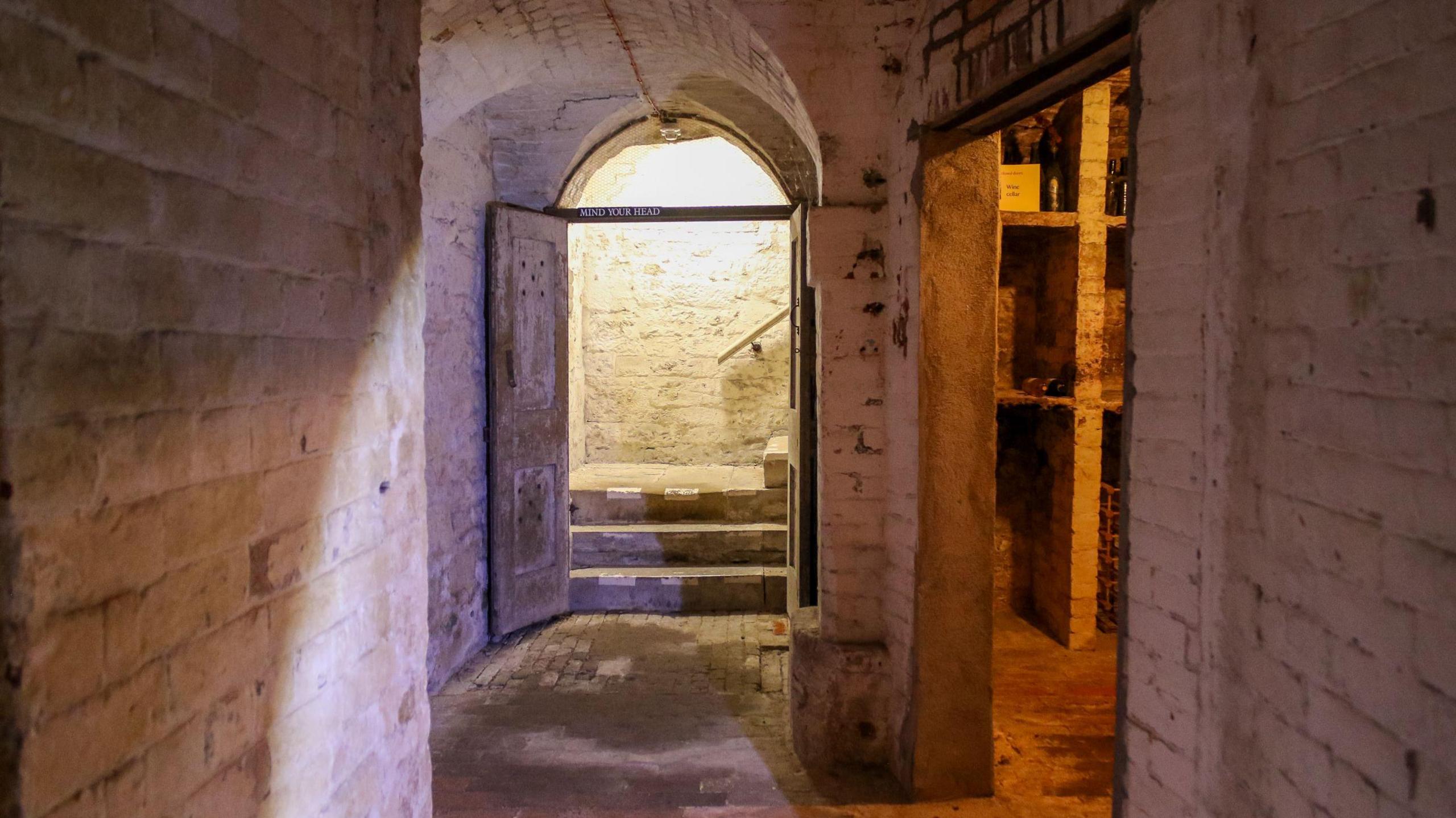 an underground passage with a door and steps leading upwards in the distance. To the right, another dimly-lit room with old-fashioned shelves