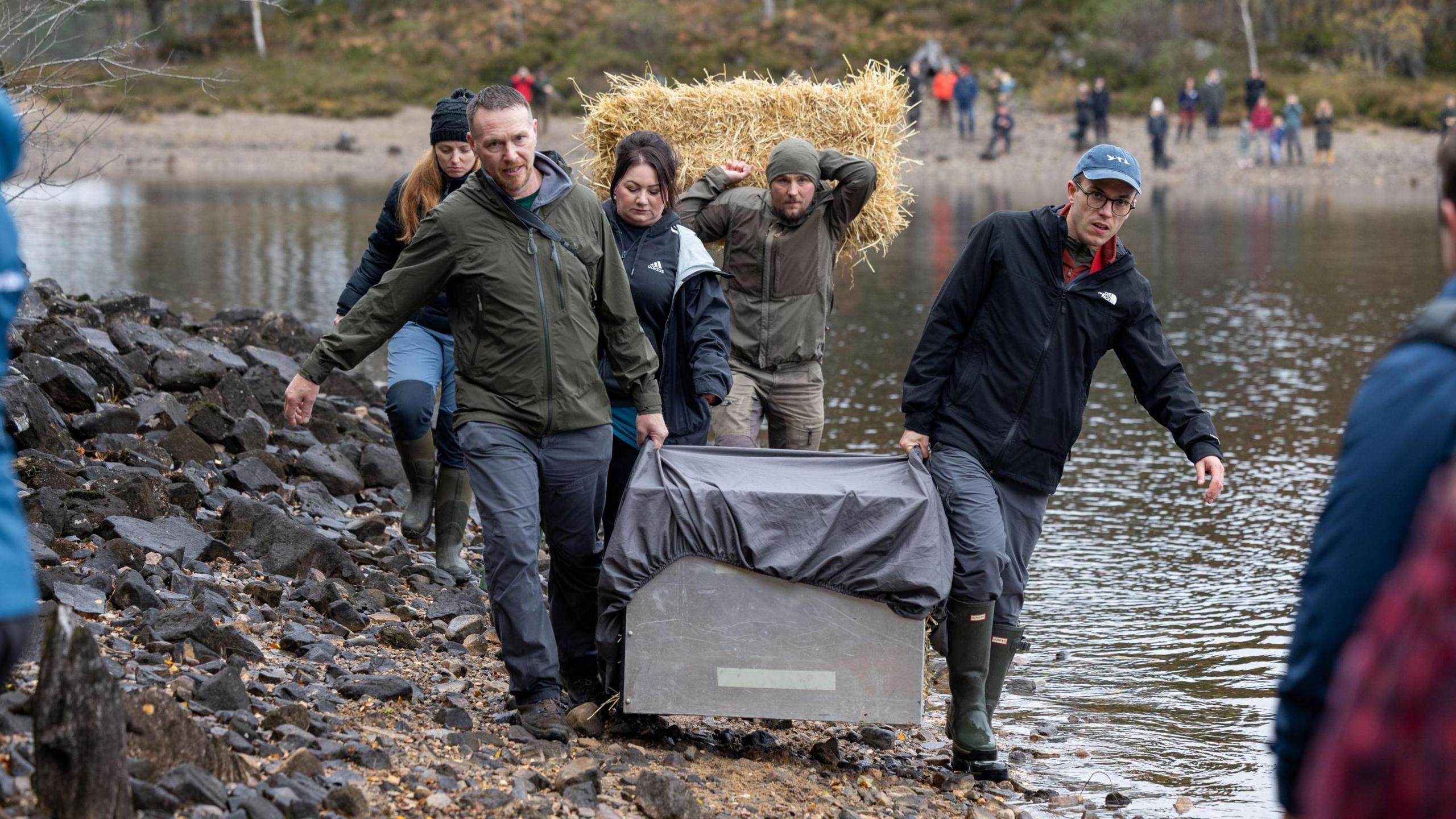 A group of people dressed in outdoor clothing walk together along a shore of the loch. Two men carry a large metal crate. A man behind them carries a bale of straw.